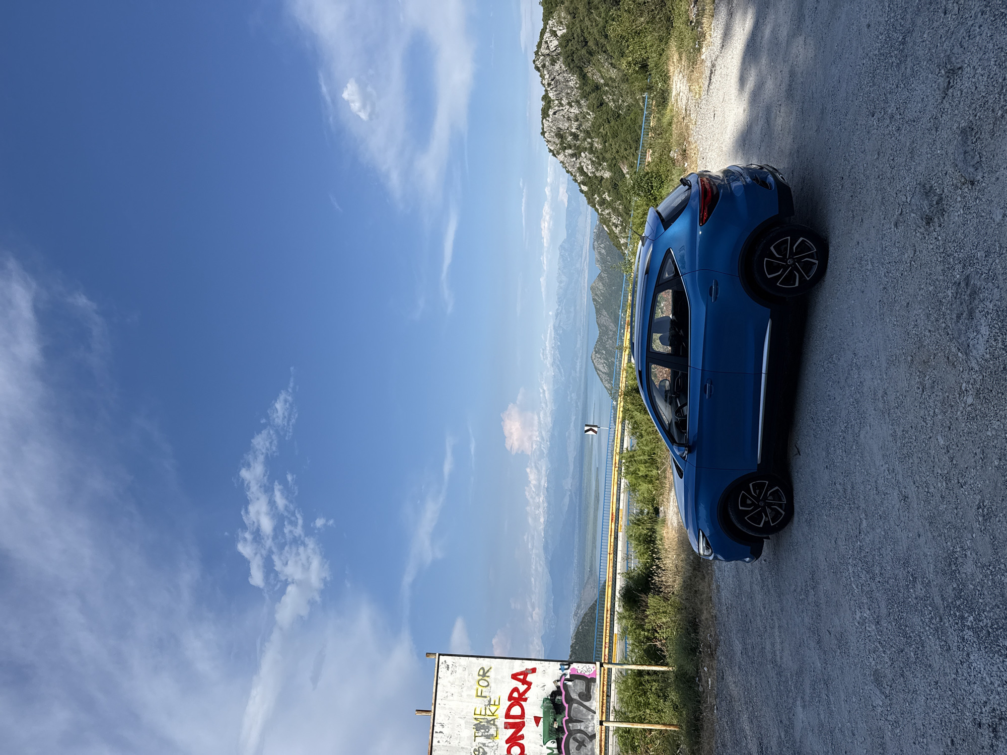 A bright blue car is parked on a gravel road overlooking a vast, mountainous landscape with a body of water in the distance. The car is positioned on the right side of the frame, facing away from the viewer. Its wheels and part of its side profile are visible.
The scene is set during the daytime, with a clear blue sky dotted with white clouds. The sun is shining, casting light on the car and the surrounding environment. To the left of the car, there is a steep, rocky mountain covered in greenery. In the background, a hazy, expansive view of mountains and a large body of water unfolds, suggesting a coastal or lake region.
In the foreground, on the lower left side of the image, a sign with graffiti is visible. Parts of the text on the sign are legible, including "IT'S TIME FOR" and "ONDRA," along with some abstract graffiti art in bright colors. A yellow barrier fence runs along the edge of the road, separating it from the drop-off with the view. The overall impression is one of a scenic overlook, possibly a viewpoint or a stop along a mountain road. The setting appears serene and offers a panoramic vista.