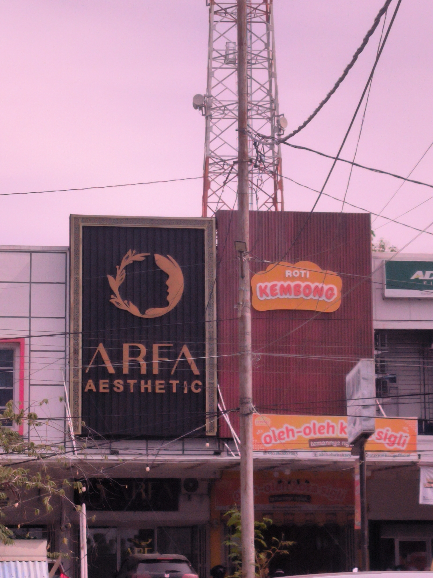 This eye-level, medium shot captures a bustling commercial street scene in Cot Teungoh, Indonesia. The sky, a soft pinkish-purple, suggests either early morning or late afternoon, or a prominent color cast. A tall, industrial communications tower dominates the right side of the background, its metal lattice structure extending high into the diffuse sky. Numerous overhead utility wires and cables crisscross the frame, connecting to a prominent utility pole in the mid-ground and partially obscuring the buildings.
Several commercial storefronts line the street. On the left, a large black rectangular sign with gold accents advertises "AREA AESTHETIC," featuring a distinctive logo of a stylized face profile encased within a leafy wreath. Adjacent to it, a building with a dark red corrugated metal facade displays a prominent orange, cloud-shaped sign that reads "ROTI KEMBONG" (Puffy Bread). Below this, another orange sign, partially obscured by the pole and wires, advertises "oleh-oleh k sigli" (souvenirs from Sigli). Smaller text below includes "temannya ng," "abibababakery," and "otikemhongaf," alongside a phone number: "57085360007085." Further right, a small green sign shows "AD." Faintly visible below is additional text: "1-oleflota sigti."
Green tree branches are visible in the lower left foreground, and the top of a dark-colored car is partially visible at the bottom of the frame. The scene is static, depicting an urban environment characterized by commercial activity and extensive overhead infrastructure.