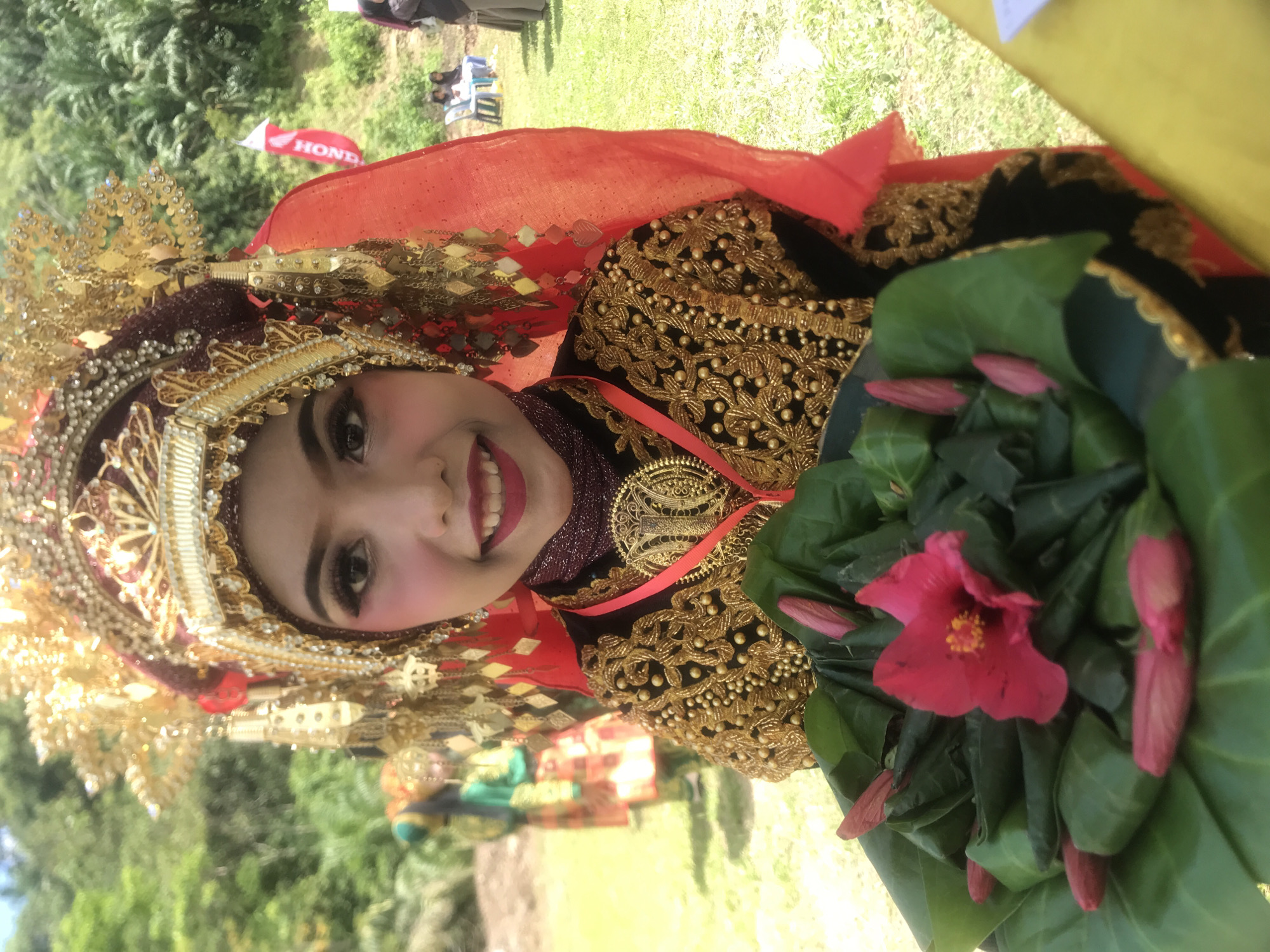 A young person in traditional Indonesian attire, specifically a rich, embroidered garment with gold embellishments and a large, ornate headdress adorned with gold and red accents, smiles at the camera. They hold a bowl filled with green leaves and pink flowers. The background shows a grassy area with trees and hints of distant structures and people, suggesting an outdoor event. A red flag with the word "HONDA" is visible on the right. The overall scene appears to be a cultural celebration or performance in Langkuta, Indonesia, given the traditional dress and festive atmosphere.