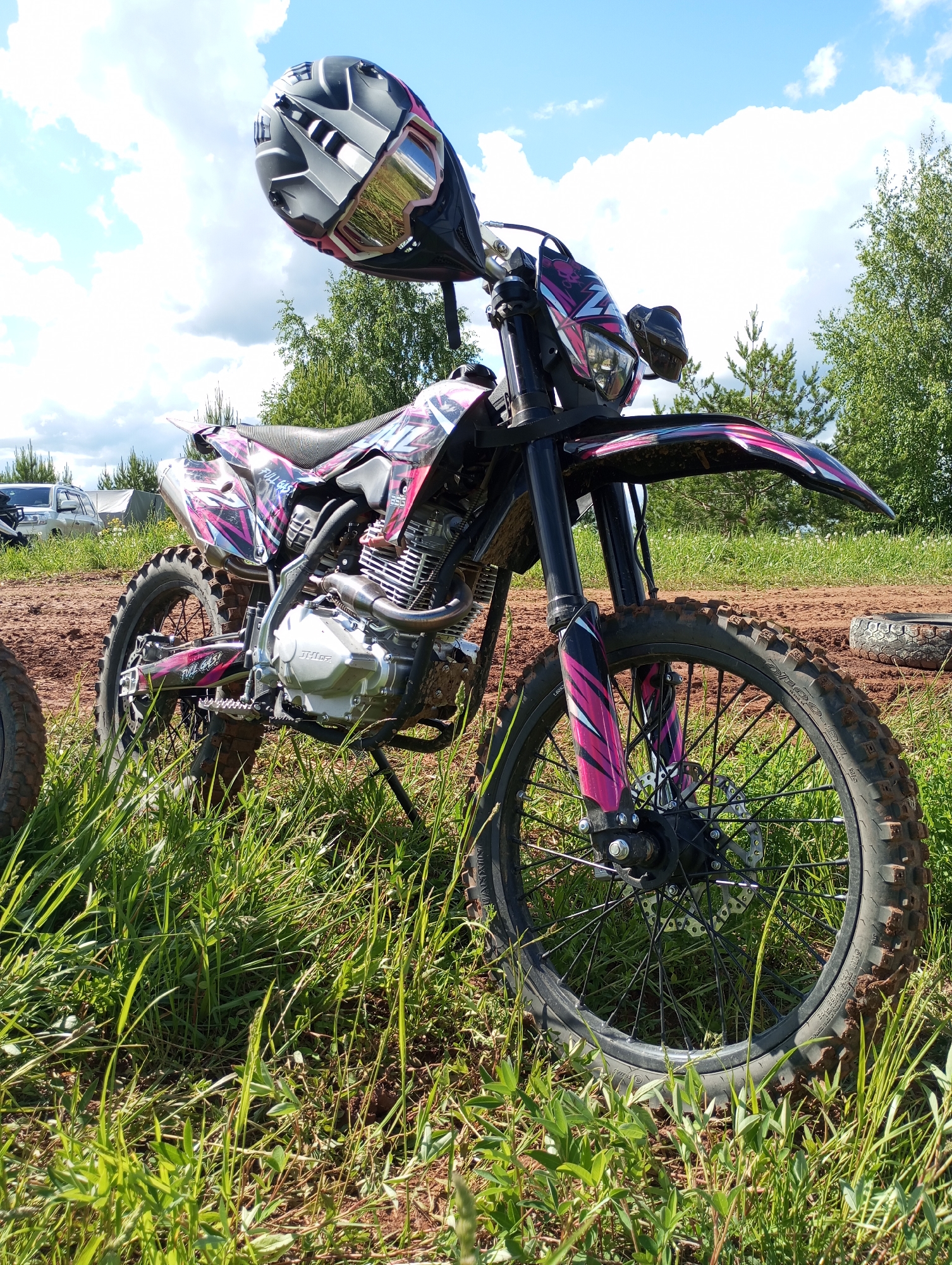 The image shows a pink and black dirt bike parked on a grassy area with a dirt road in the background. The bike is angled towards the viewer and a helmet rests on the handlebars. The surrounding environment includes green grass, trees, a cloudy sky, and in the distance, other vehicles and possibly tents are visible. The location of Izhevsk, Russia is not confirmed.
