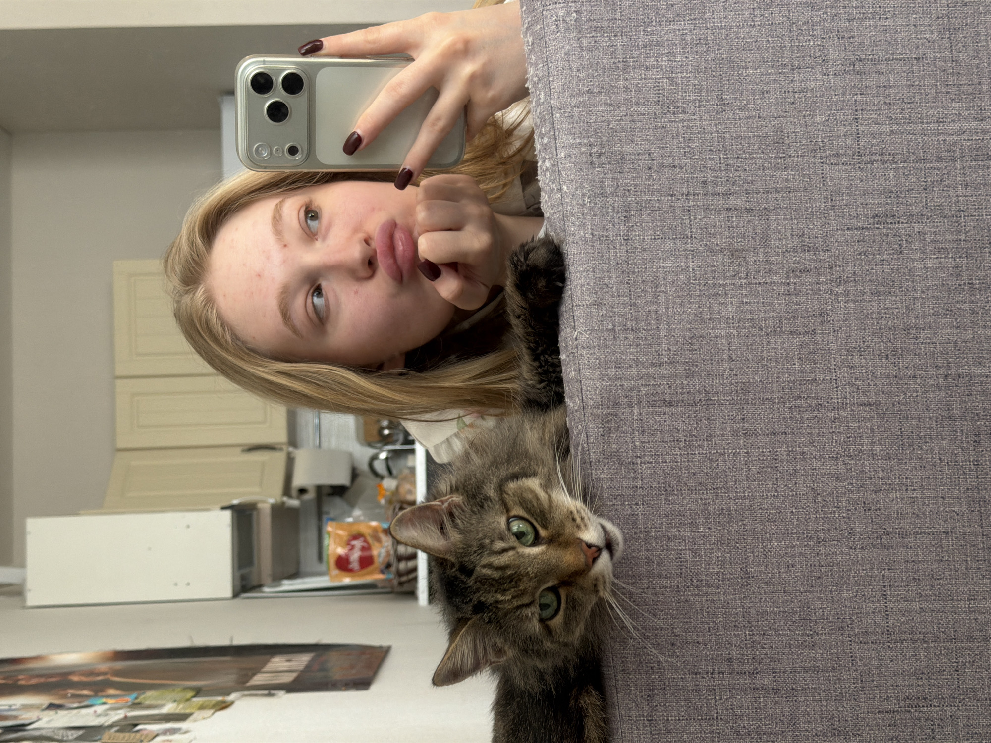 A young woman is holding a smartphone to take a selfie with a tabby cat. The woman is pouting at the camera and holding up her hand, which has dark red nail polish. The cat is looking directly at the camera. They are positioned behind a gray sofa. In the background, there's a glimpse of a kitchen area with cabinets. The location is Засечное, Russia.