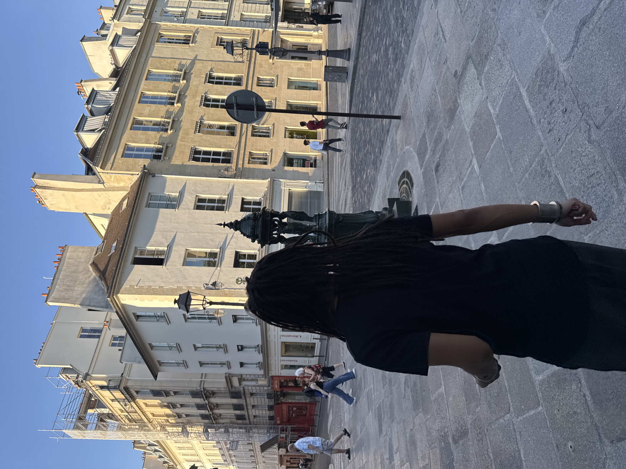 The image depicts a person with dreadlocks, facing away from the camera, standing on a cobblestone square in Paris, France. The person is wearing a black shirt and pants and has a metal bracelet on their wrist. In the background are Parisian buildings with multiple windows and architectural details. There is a Wallace fountain to the left of the person and a street sign is in the distance, with a couple of people walking by it. To the left is scaffolding. Other pedestrians are scattered throughout the scene. The sky is a clear blue. The cobblestone square is composed of grey stones.