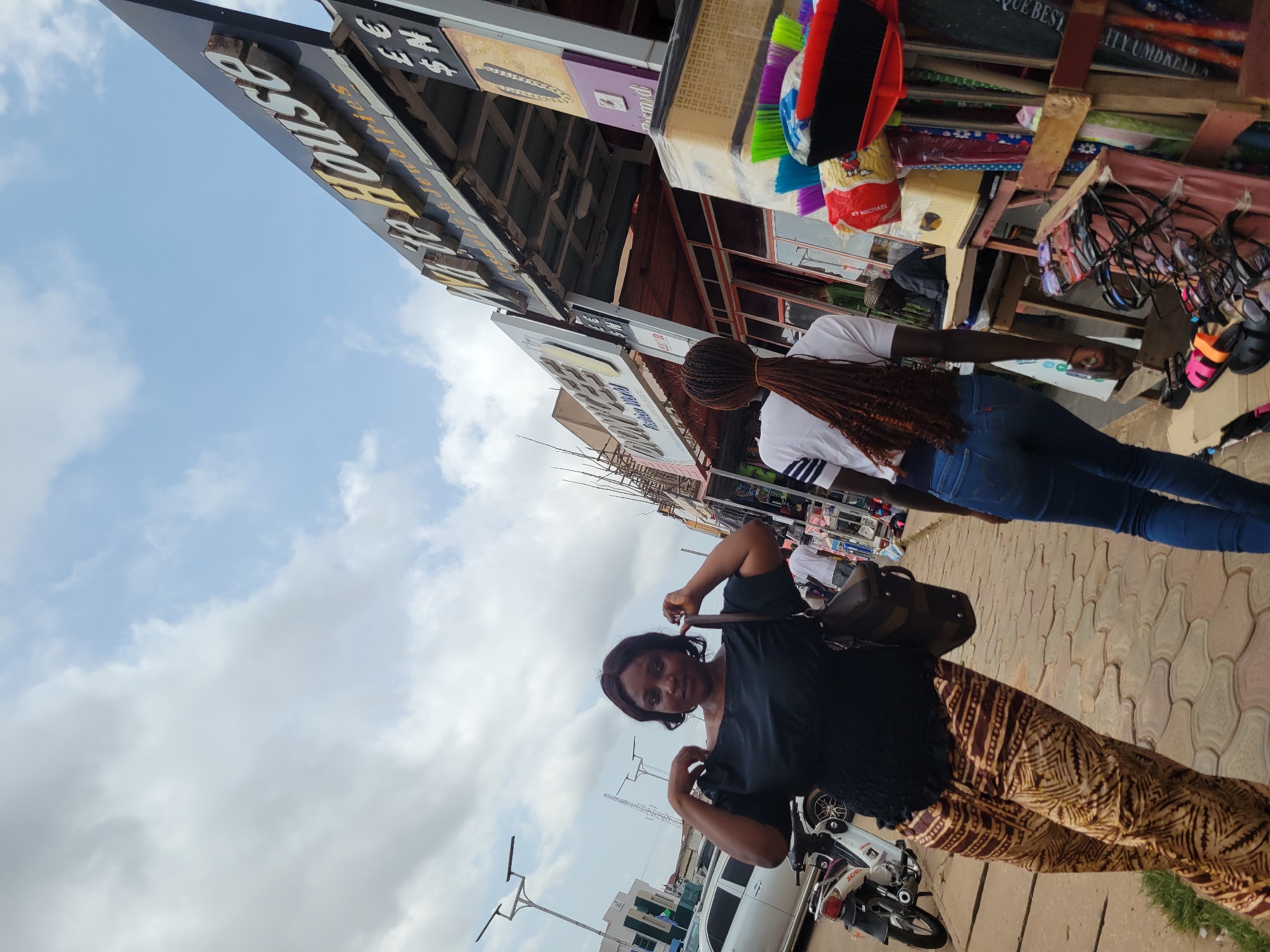 The image captures a street scene in Jos, Nigeria, likely a commercial area. In the foreground, two women are walking along a paved sidewalk. The woman on the left, facing the camera, is wearing a black off-the-shoulder top and patterned trousers, and is holding a brown handbag. The woman on the right, with her back to the camera, is wearing a white t-shirt with blue stripes and blue jeans, her long braided hair extending down her back.
The background features a row of shops. A prominent sign reads "White House" with "Cosmetic & Jewelries" below it, and currency symbols like €, £, and $N are visible. Another sign to the left reads "DONCEE Fashion World". Various items are displayed outside the shops, including brooms, brushes, and what appear to be sunglasses and flip-flops. A motorcycle is parked on the left side of the street, and a silver car is visible further down. The sky is overcast with clouds. The overall impression is that of a bustling market or shopping street.