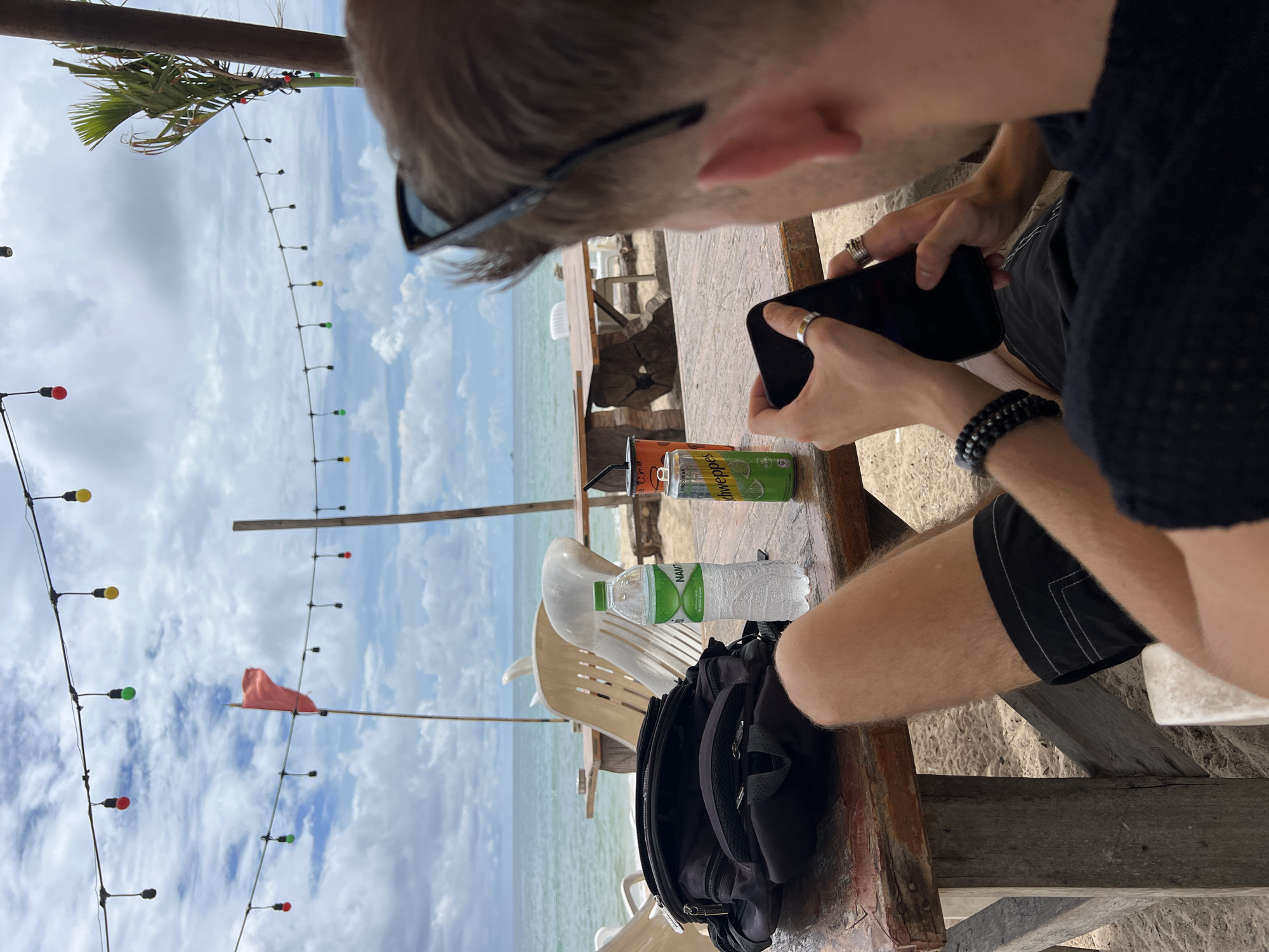 In an outdoor setting on a sandy beach, a person is seated at a wooden table, looking down at their smartphone. The person is wearing a black t-shirt and black shorts with white stitching. They have several rings on their fingers and a beaded bracelet on their left wrist. On the table are a black backpack, a bottle of water, and two cans of soda. In the background, there is the ocean, chairs, and strings of colorful lights and a red flag. The sky is partly cloudy.