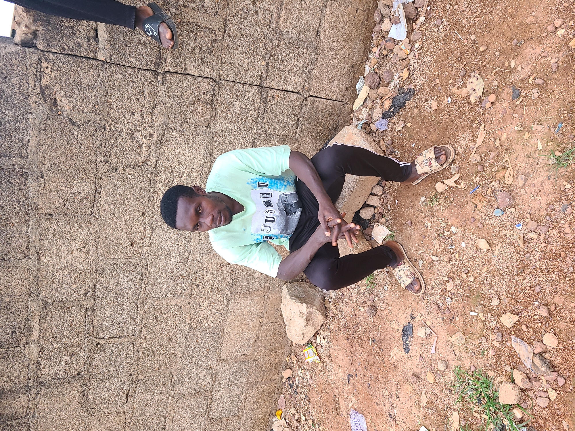 A man is sitting on a rock with his hands clasped in front of him, leaning against a rough, grey stone wall. He is wearing a light green t-shirt with a graphic print and black pants, and brown sandals. The ground in front of him is reddish-brown dirt scattered with small rocks, debris, and some white and yellow wrappers. To the right of the man, another person's legs are visible, wearing black pants and black flip-flops with a white Nike logo. The overall environment suggests an outdoor, urban setting in Jos, Nigeria, characterized by its unpaved ground and simple, functional architecture.