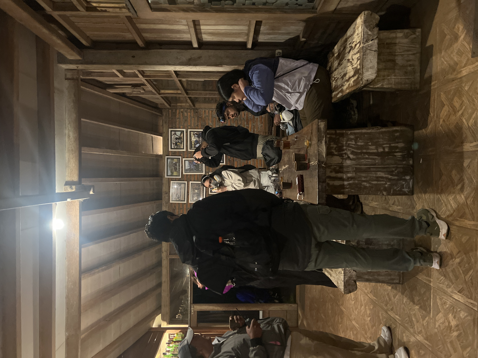 The image shows a group of people inside a wooden building, likely a restaurant or gathering place, in Unknown, Indonesia. The interior features exposed wooden beams for the ceiling and a wooden floor. People are standing around a wooden table, with some appearing to be taking photos with cameras. The walls are adorned with framed pictures. There are wooden benches or stools for seating. Bottles are visible on a shelf in the background.