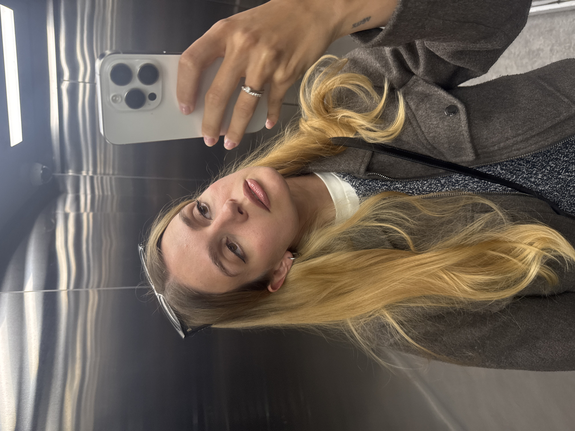 A young woman is captured in a mirror selfie, holding a white smartphone with her left hand. Her blonde hair cascades over her shoulders, and she wears a dark gray textured coat over a white top and a dark, speckled garment. A silver-toned ring is visible on her left ring finger, and a small tattoo, possibly reading "ARTEM", is on her left wrist. She has winged eyeliner, light-colored lipstick, and a small hoop earring on her right ear. A pair of glasses is pushed up onto her head.
The woman's gaze is directed towards the camera with a calm expression and a slight pout. The smartphone she is holding appears to be a modern iPhone, identifiable by its distinct triple-camera module.
The background consists of highly reflective, vertically-brushed metallic walls, strongly suggesting an elevator interior. Bright, vertical light sources are prominently reflected on the glossy surface, creating dynamic patterns of light and shadow on the metallic surface.