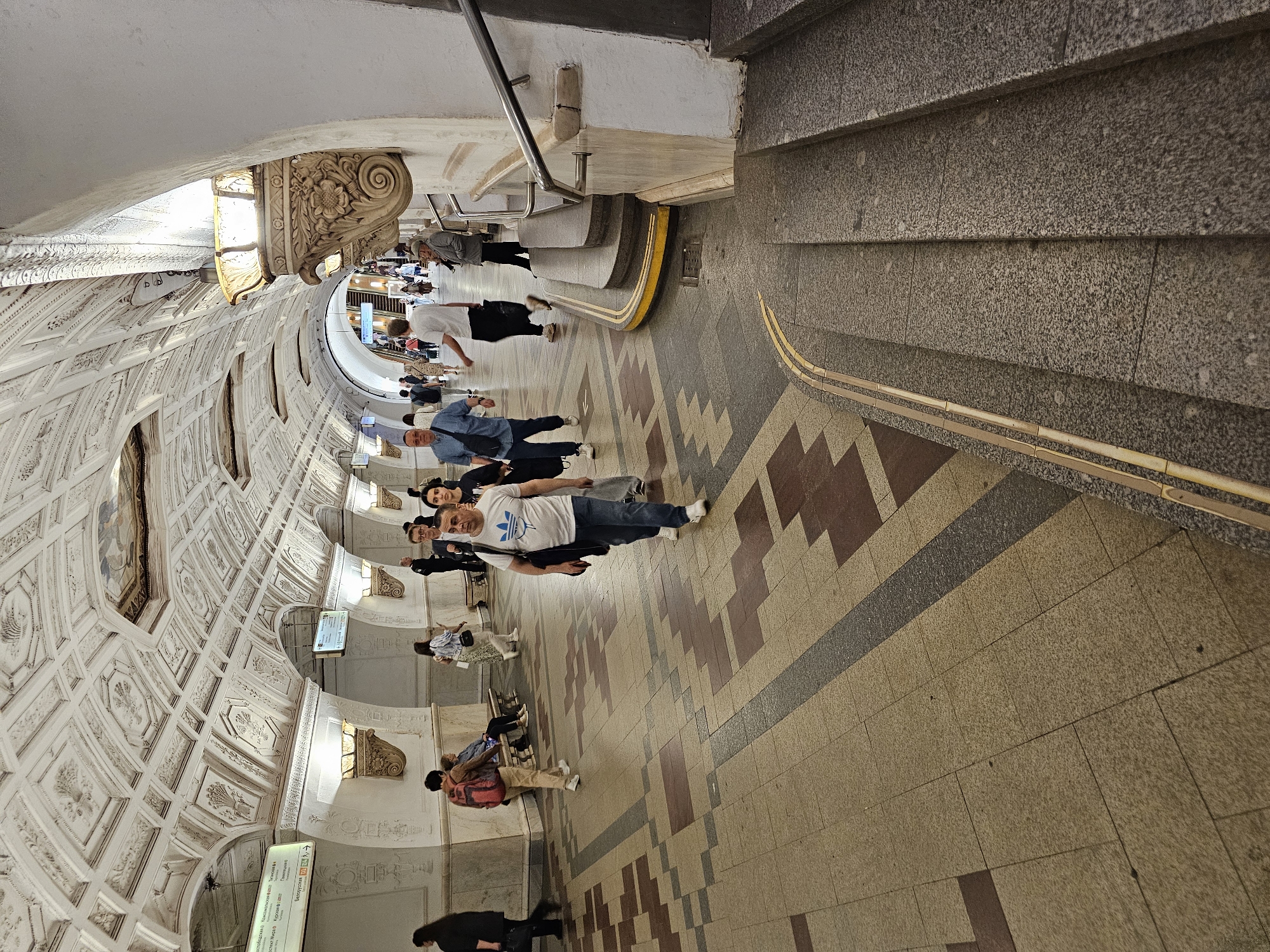 This is a photograph of the Kievskaya Metro Station in Moscow, Russia. It shows an elaborately decorated hallway with a vaulted ceiling featuring intricate plasterwork and arches. A group of people are walking along the patterned tiled floor of the corridor. There are stairs on the right side of the image with a metal railing, and a tunnel-like opening at the far end of the hallway, with additional people visible within. The scene is well-lit.