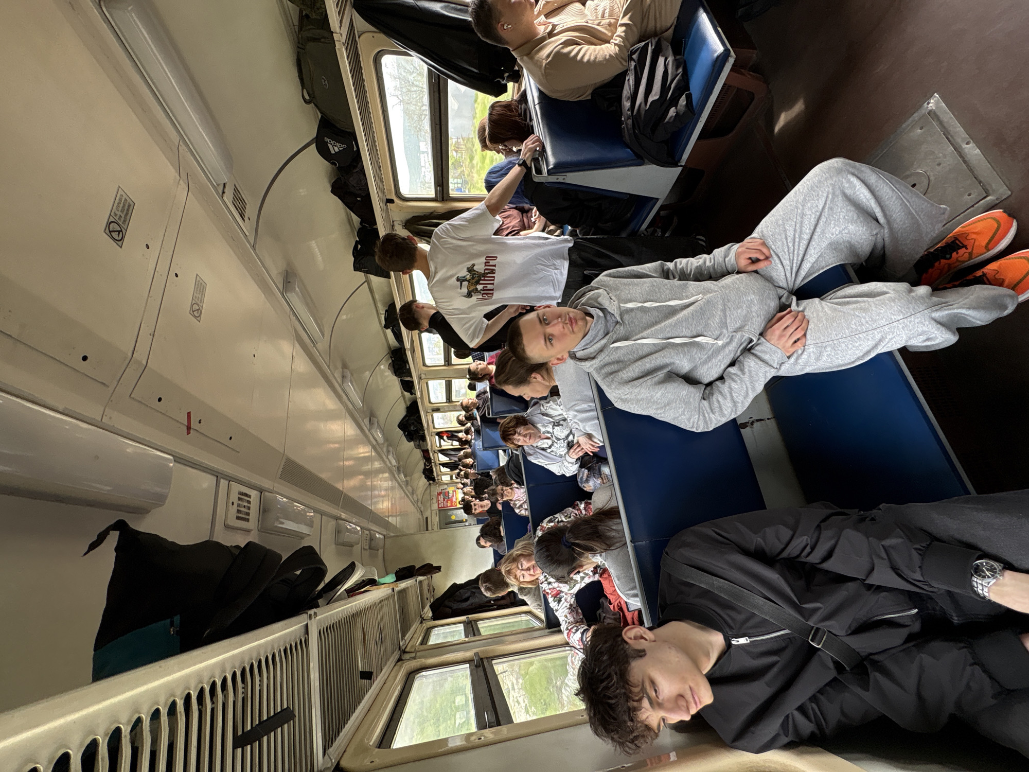 The image depicts the interior of a train carriage filled with passengers. The passengers are seated, some are sleeping, and others are looking around. Overhead luggage racks are visible, filled with various bags and personal items. The train car appears to be well-lit. It is not possible to determine from the image if this scene takes place in the city of Kirillovka, Russia, or another location.