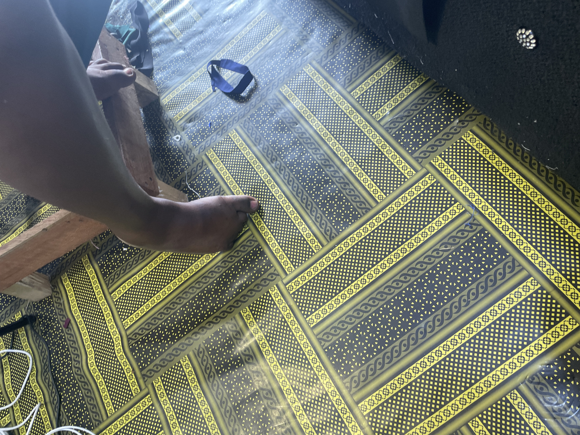 An overhead shot shows a person's bare foot resting on a floor covered in a dark, patterned linoleum. The linoleum has a repeating design of yellow bands with intricate geometric and wavy motifs. The person's leg is visible, extending from the top right of the frame, and their foot appears to be placed on or near a wooden structure. A dark blue elastic band lies on the floor to the right of the foot. The overall lighting suggests an indoor setting.