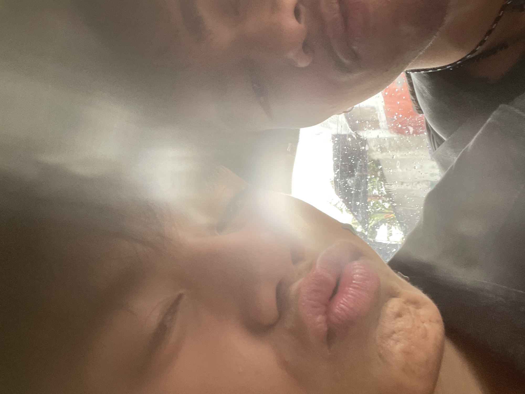 The image is a close-up selfie of two individuals, likely taken inside a vehicle. The person on the left is facing the camera with their eyes closed and lips puckered in a kissing gesture. Their skin appears to have some texture on the chin. The person on the right is partially visible, with their face turned slightly away from the camera. A necklace with a patterned string is visible around their neck. Through the window behind them, a blurred background suggests an outdoor environment with some greenery and what appear to be buildings. The presence of raindrops on the window indicates it might be raining. Without further context or visual cues, determining the precise location within Davao City, Philippines, is not possible from the image alone.