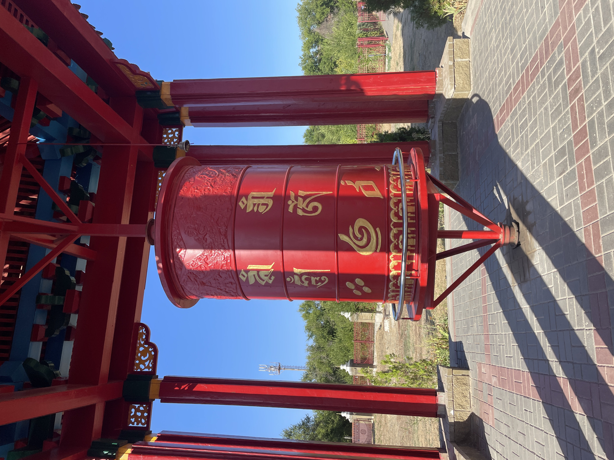 The image shows a red Buddhist prayer wheel in Elista, Russia. The prayer wheel is cylindrical and decorated with golden Buddhist inscriptions. It is housed under a red pagoda-style roof supported by red pillars. The ground is paved with gray and red interlocking bricks, and shadows from the structure are cast across the paving. In the background, there are green trees, a fence, and a cell tower. The sky is clear blue. There are no people in the image.