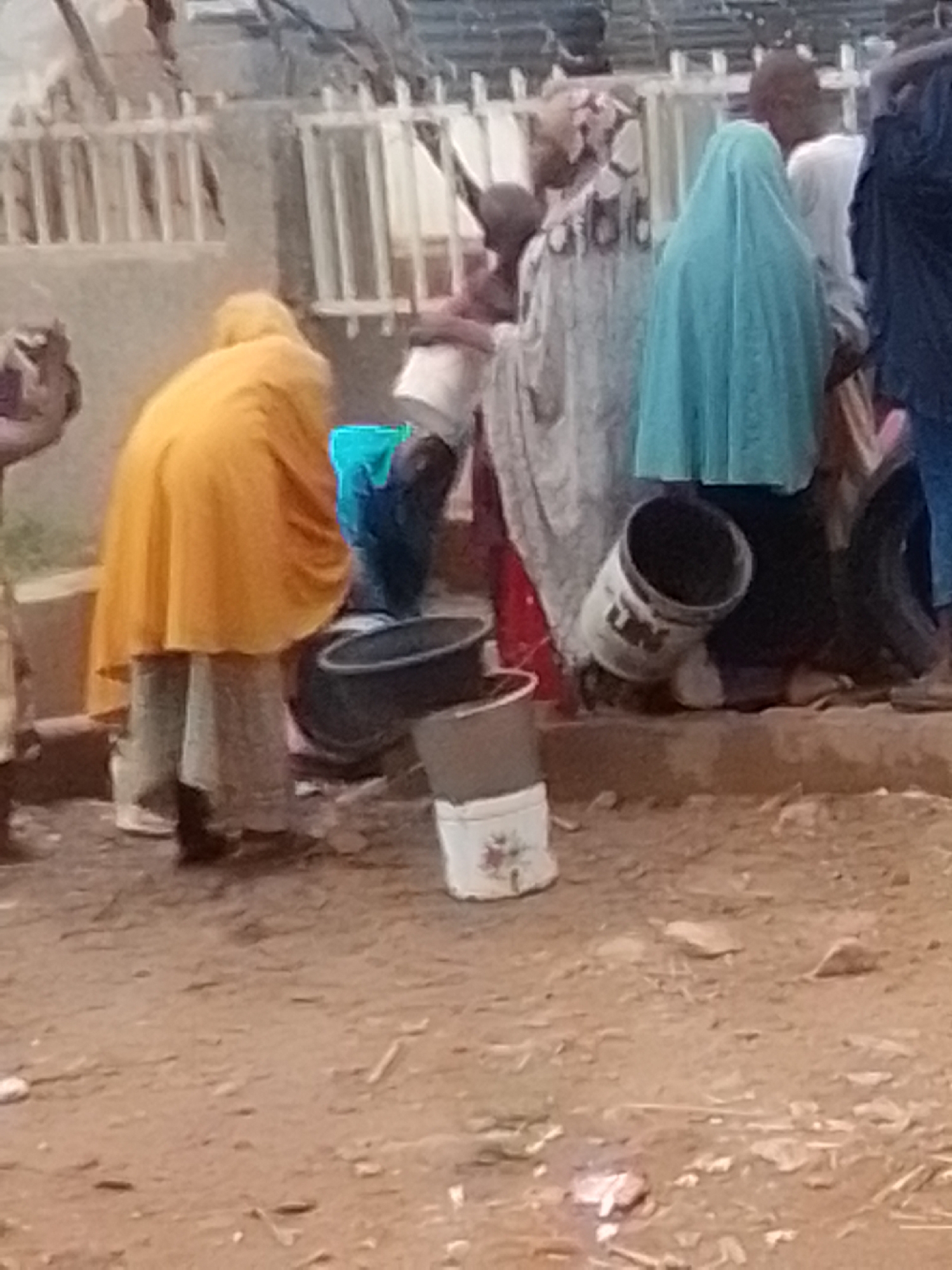 The image depicts a dusty, outdoor scene in Pantame, Nigeria, where several individuals are gathered. In the foreground, a person wearing an orange garment and light-colored trousers is bent over, seemingly tending to something near large buckets. To their right, a person in a light blue headscarf is also bent over, facing away from the camera. Behind them, a person in a white garment is partially visible, and further back, another individual in dark clothing stands with their back to the viewer. In the background, structures with white railings and debris are visible, suggesting a potentially impoverished or developing area. The ground is covered in dirt and scattered debris, with several buckets and containers present, including a white bucket with floral decorations. The overall impression is one of communal activity, possibly related to water collection or domestic chores, in a gritty urban environment.