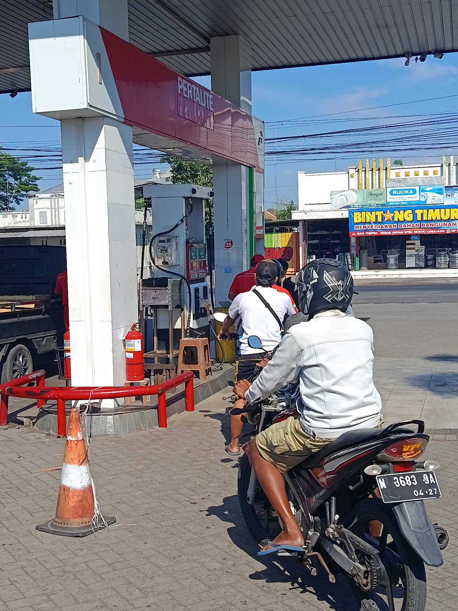 The image shows a view from behind a motorcycle at a gas station in Tempeh Lor, Indonesia. A person is sitting on the motorcycle, wearing a white jacket, khaki shorts, and a black helmet with silver accents. Another person in a white shirt and dark shorts is also on a motorcycle in front of the first. Behind them, a person in a red shirt is standing next to a pickup truck. The gas station has a red and white sign that says "PERTALITE". There are gas pumps, a fire extinguisher, and a traffic cone visible. In the background, there is a building with a sign that says "BINTANG TIMUR" and appears to be a building materials store. The sky is blue and clear.