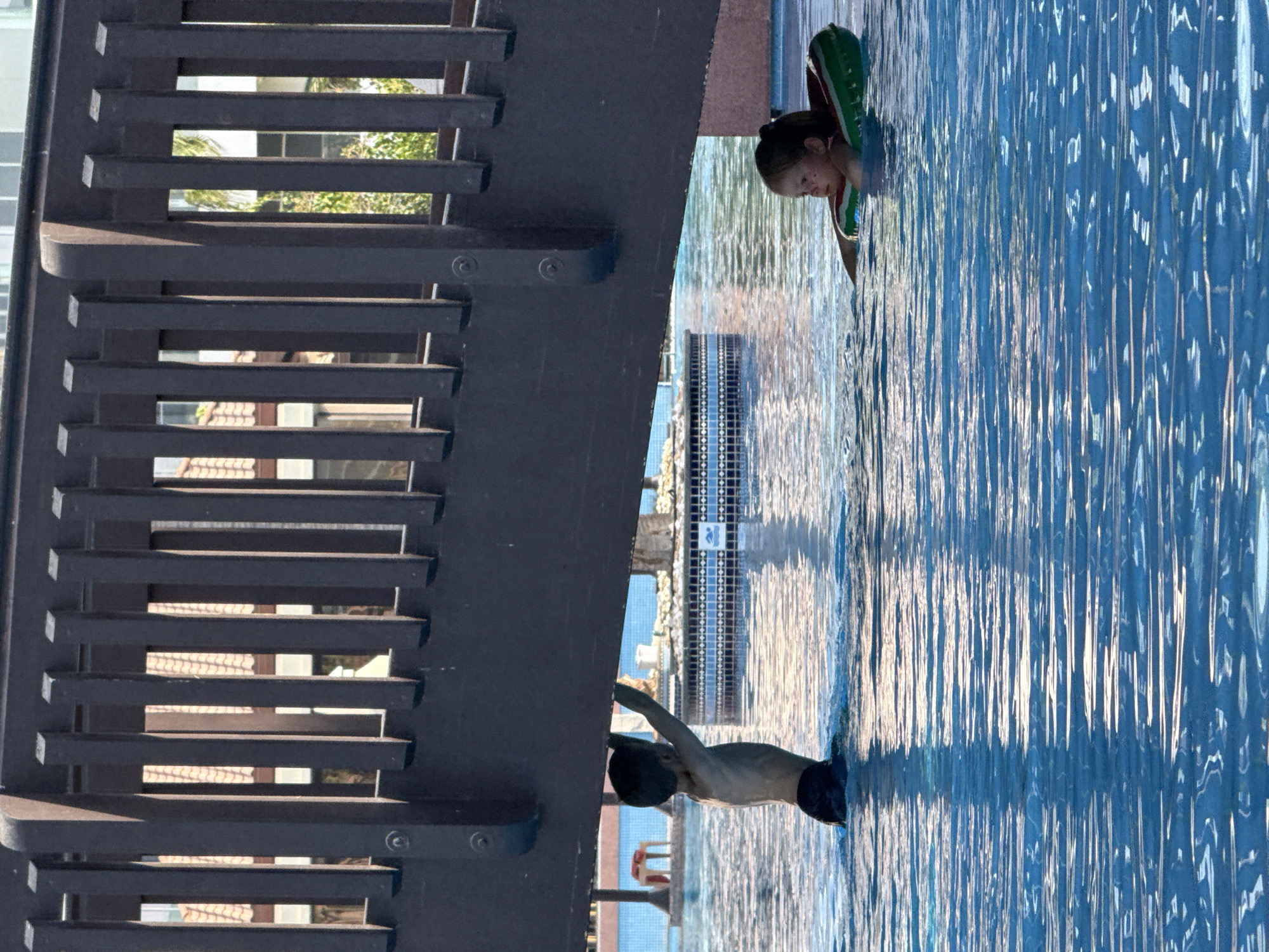 Two children are in a swimming pool under a wooden bridge in Dubai, United Arab Emirates. A young boy, with dark hair and wearing dark swim trunks, stands in the water, reaching up to touch the underside of the bridge. To his right, a young girl with dark hair, wearing a pink swimsuit and a green watermelon-shaped inflatable ring, floats in the pool. The water is bright blue and reflects sunlight, creating ripples and patterns. The bridge is made of dark wood and has a railing with vertical slats. In the background, parts of buildings with orange tiled roofs and palm trees are visible through the bridge's structure, suggesting an outdoor setting with resort-like architecture. The water in the pool appears clear and inviting.