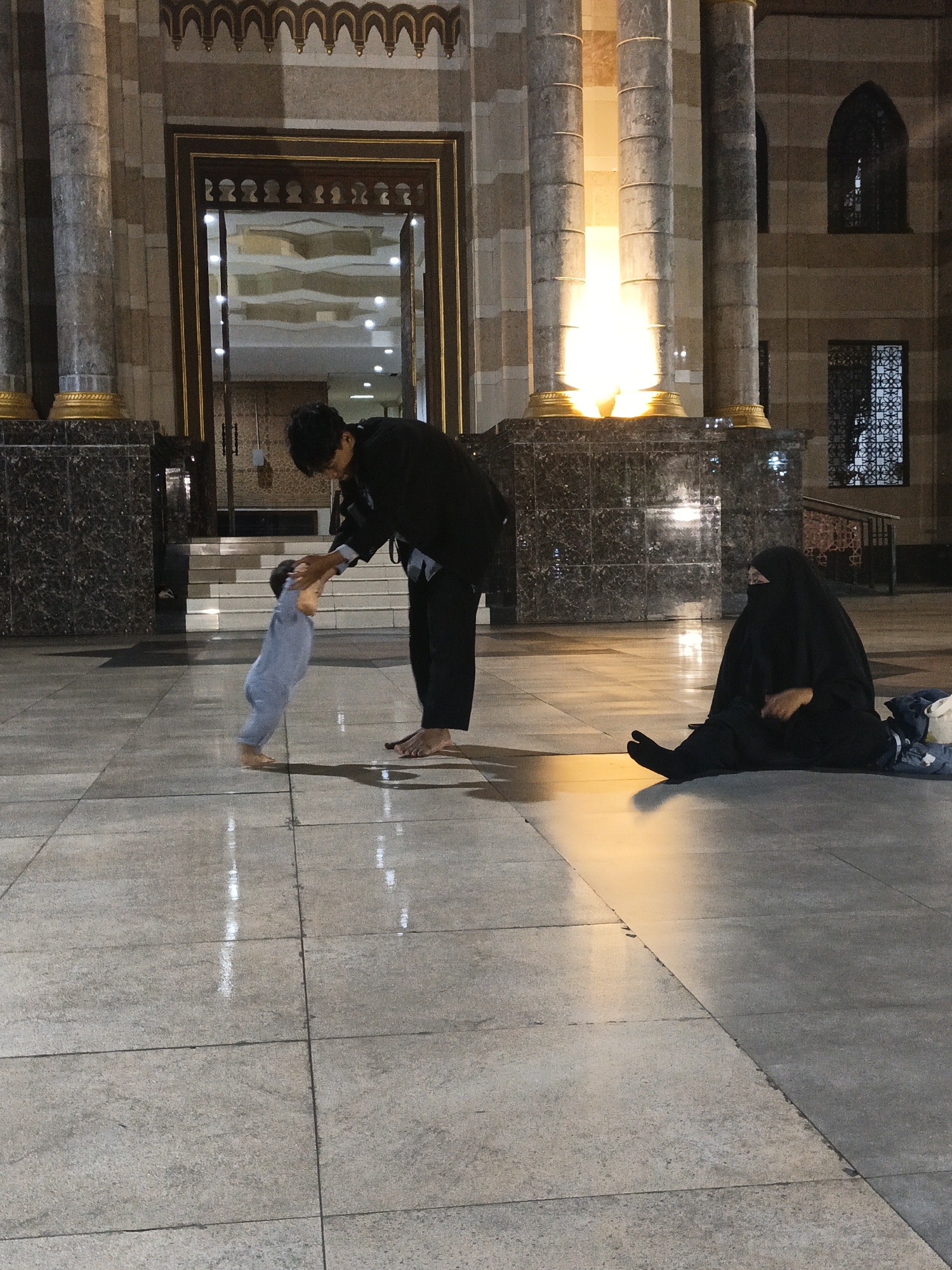 A father is teaching his baby to walk in front of a mosque in Jonggrangan, Indonesia. The father, dressed in a black shirt and pants, is holding the baby's hands as the baby takes a step forward. The baby is wearing a light blue onesie. A woman, dressed in a black hijab and abaya, is sitting on the floor to the right, facing away from the camera. The floor is made of large, grey tiles and is wet, reflecting the lights from the mosque. The mosque has tall, stone columns and ornate arched doorways. The lighting suggests it is nighttime or dusk.