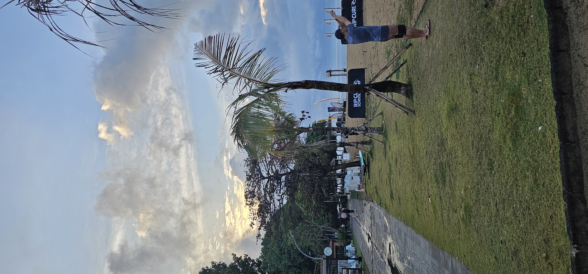 The image depicts a coastal scene in Denpasar, Indonesia. The sky is overcast with a mix of blue patches and heavy clouds. A palm tree leans significantly towards the left of the frame. In the background, a beach area is visible, with a man doing some kind of exercise. A Rip Curl advertisement is visible in the frame. A pathway runs from the bottom of the frame. The landscape has grassy areas adjacent to the walkway, along with some bushes and plants. The photo seems to have been taken at an unusual angle, possibly rotated.