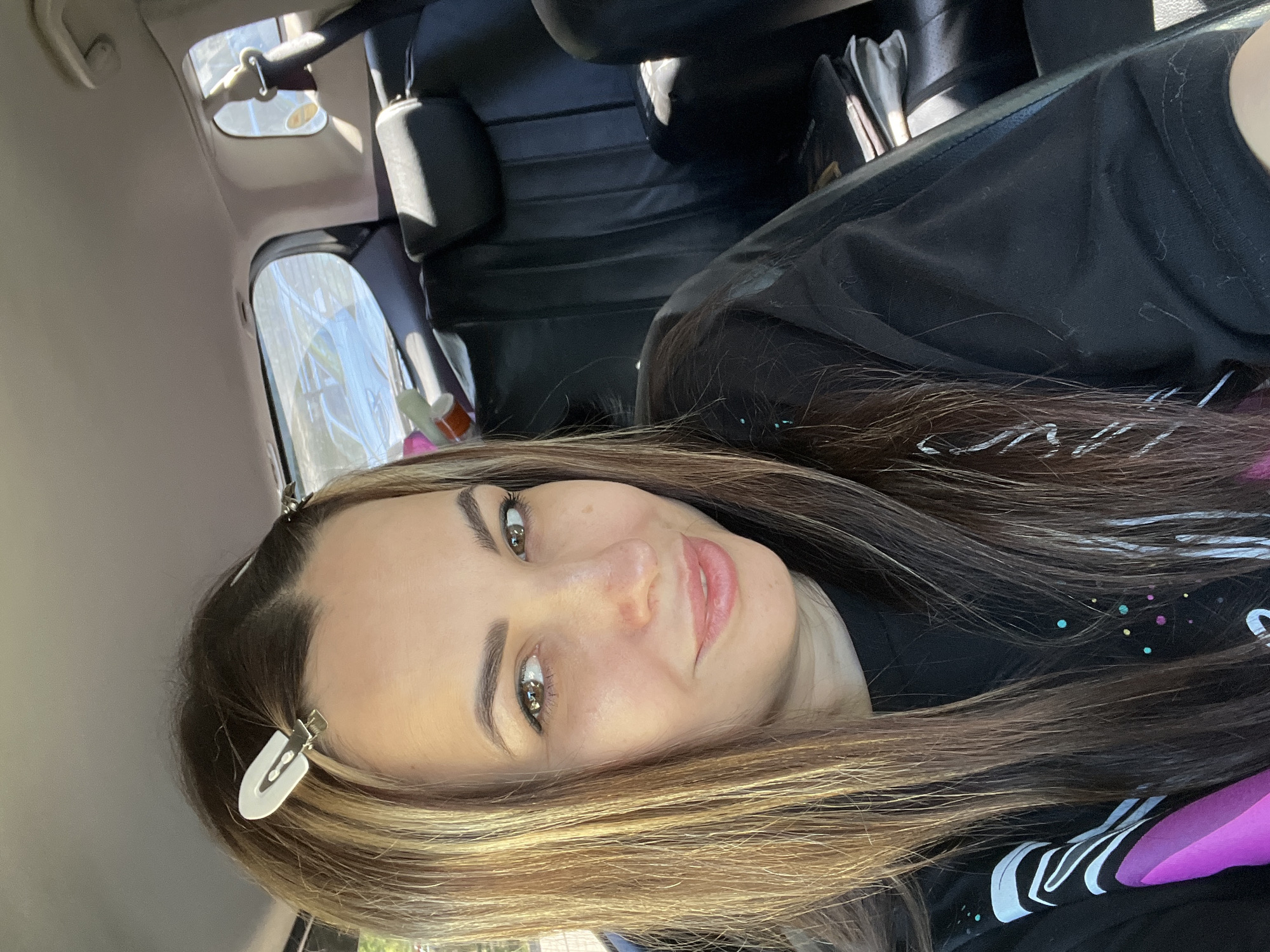 A woman with long brown hair is seated in a vehicle. She has white clips in her hair and is wearing a black shirt with a colorful design. The vehicle has black leather seats. The location is Unknown, Unknown.