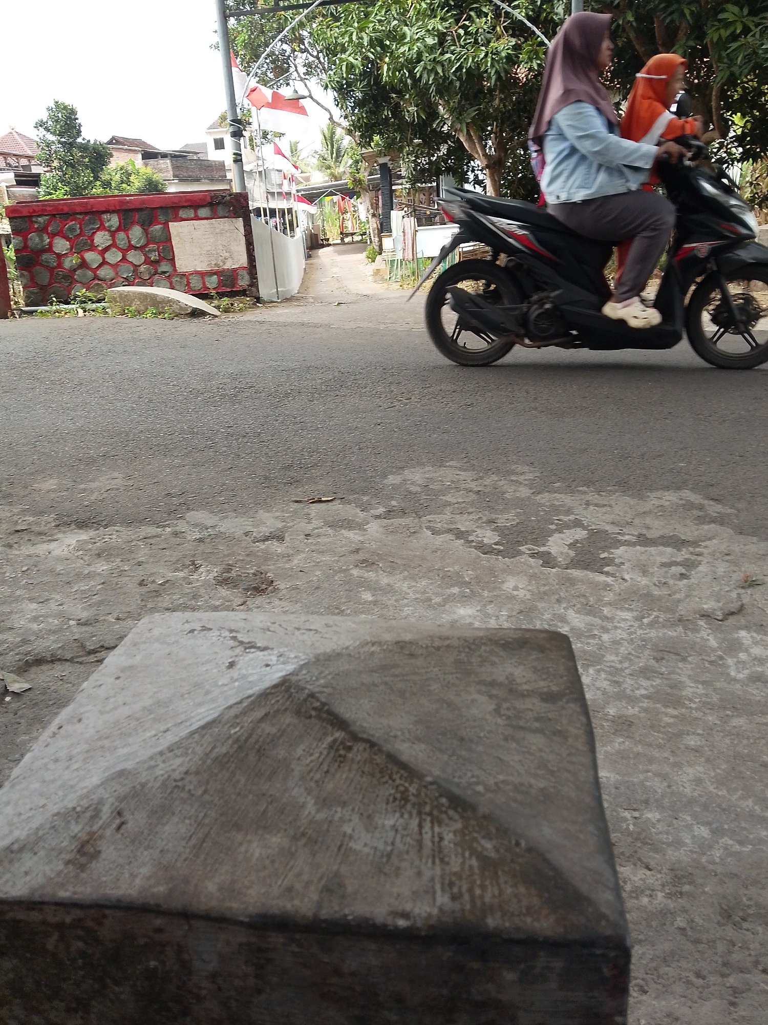In Tasikmadu, Indonesia, an image captures a snapshot of a street scene. In the foreground, a weathered concrete pillar with a pyramidal top sits on a patchy asphalt surface. The midground shows a black motorcycle in motion, carrying two individuals. The rider, a woman wearing a light blue denim jacket and a purple hijab, is behind the handlebars. A child, dressed in an orange hijab and clothing, is seated in front of her. Both are looking forward, as if traveling. In the background, a wall with a red stone facade lines the side of the road. Beyond the wall, houses with tiled roofs and lush green trees are visible. Two Indonesian flags, red and white, are displayed on a pole, adding a touch of national pride to the environment. The overall scene depicts a typical day in a residential area of the city.