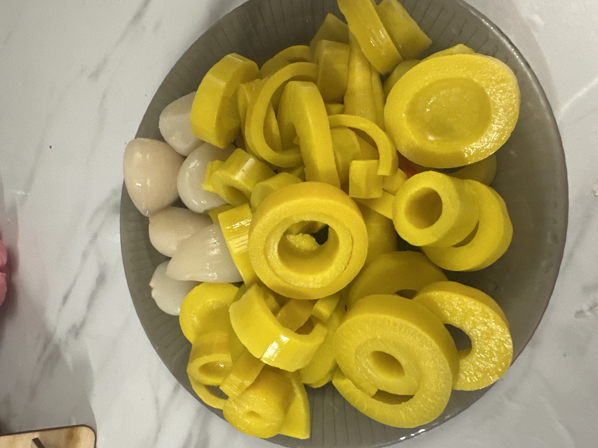 A gray plate filled with sliced yellow bell peppers and lychees is presented on a marble countertop. The bell peppers are cut into rings and some pieces are further chopped. The lychees are whole and have a translucent, white appearance. The lighting suggests an indoor setting. The image is a close-up shot focusing on the food. There is no indication of the city of Verkhniy Yurt, Russia in the image.