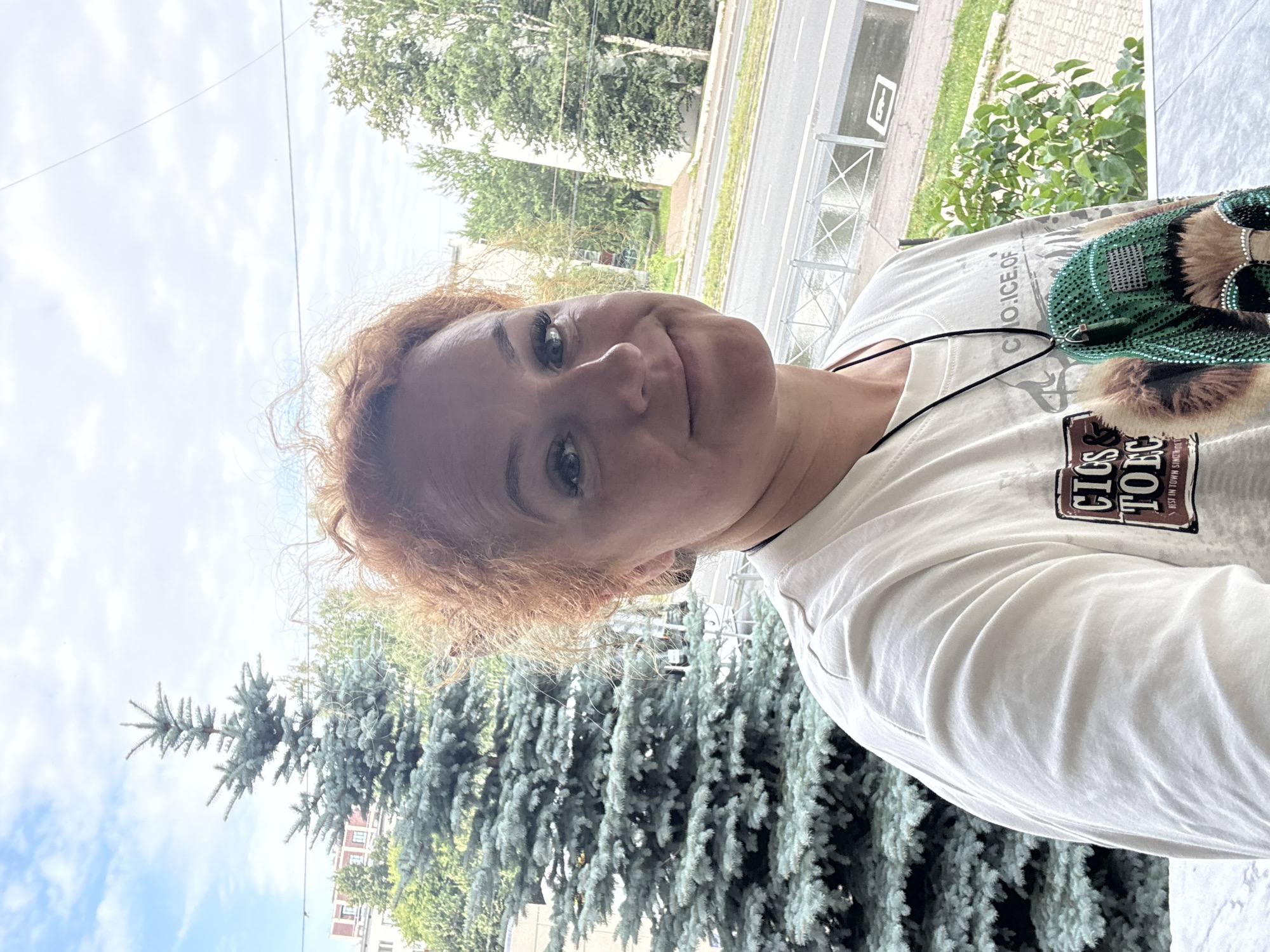 A woman with reddish-brown curly hair is taking a selfie outdoors. She is wearing a white t-shirt with a graphic print of a dog wearing sunglasses. She has on a necklace with a green pendant decorated with rhinestones and fur. Behind her, a large evergreen tree is visible, along with other trees and buildings. A street with a sidewalk and railing is in the background, suggesting an urban setting. The sky is cloudy. The woman is smiling and looking at the camera. The image appears to have been taken in Kazan, Russia, given the presence of Cyrillic text on her t-shirt which reads "CIGARS TOBACCO" and "BEST IN TOWN SINCE".