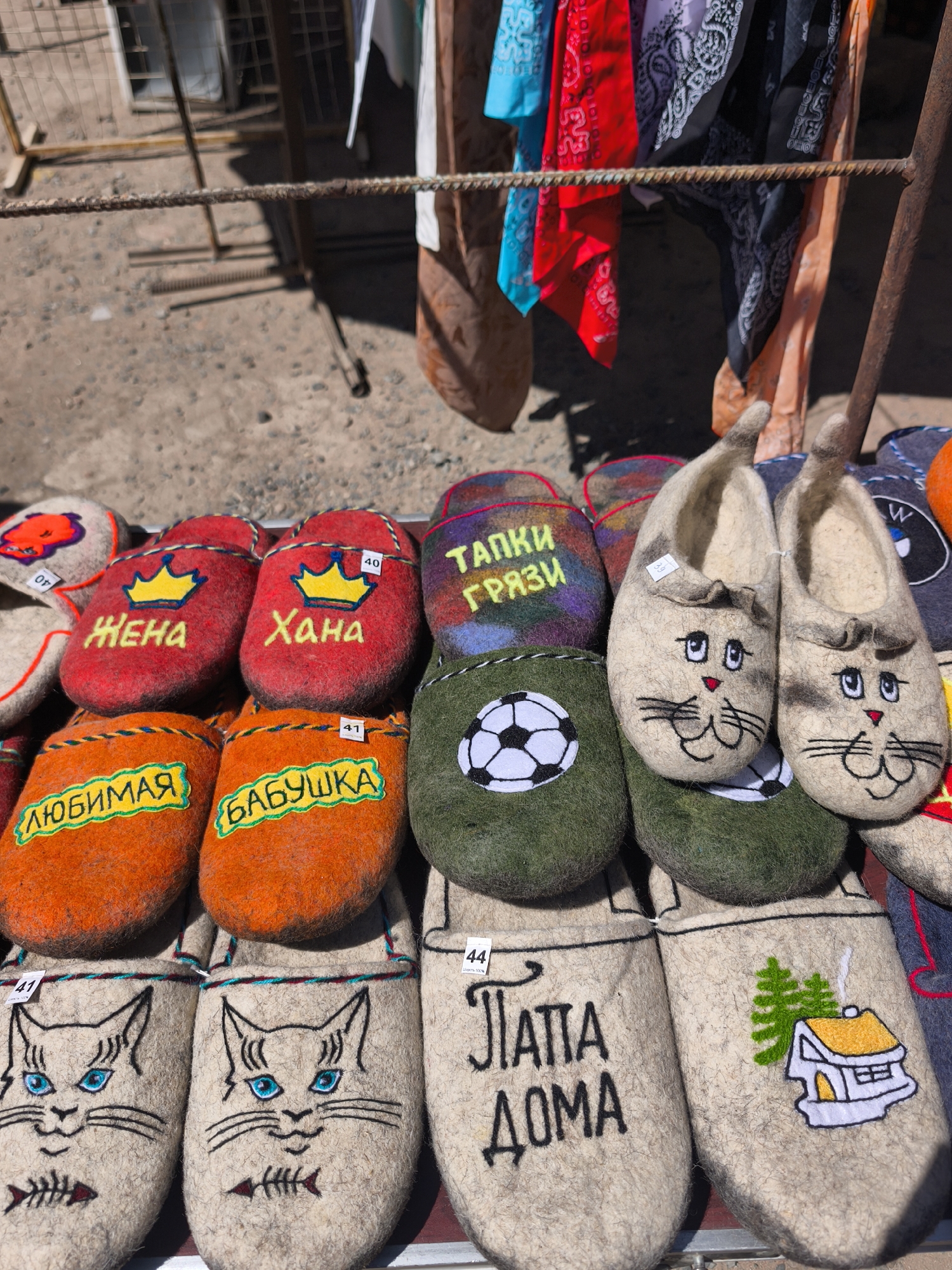 The image captures a close-up display of handmade felt slippers at a market, possibly in Bosteri, Kyrgyzstan, known for its craft traditions. The slippers are arranged in rows, each pair uniquely designed with various patterns and embroidered motifs. Some have cartoon cat faces with whiskers, while others feature embroidered words in Cyrillic, such as "ZHENA" (wife), "KHANA," "LYUBIMAYA" (beloved), "BABUSHKA" (grandmother), "TAPKI GRYAZI" (mud slippers), and "PAPA DOMA" (dad at home). One pair is decorated with a soccer ball, while another has an image of a house and a tree. Small price tags are attached to each pair. The slippers are displayed on a metal rack or table, with a rod visible in the background supporting hanging colorful textiles or bandanas. The ground appears to be pavement or concrete. The overall impression is of a vibrant and personalized selection of artisan-made slippers.