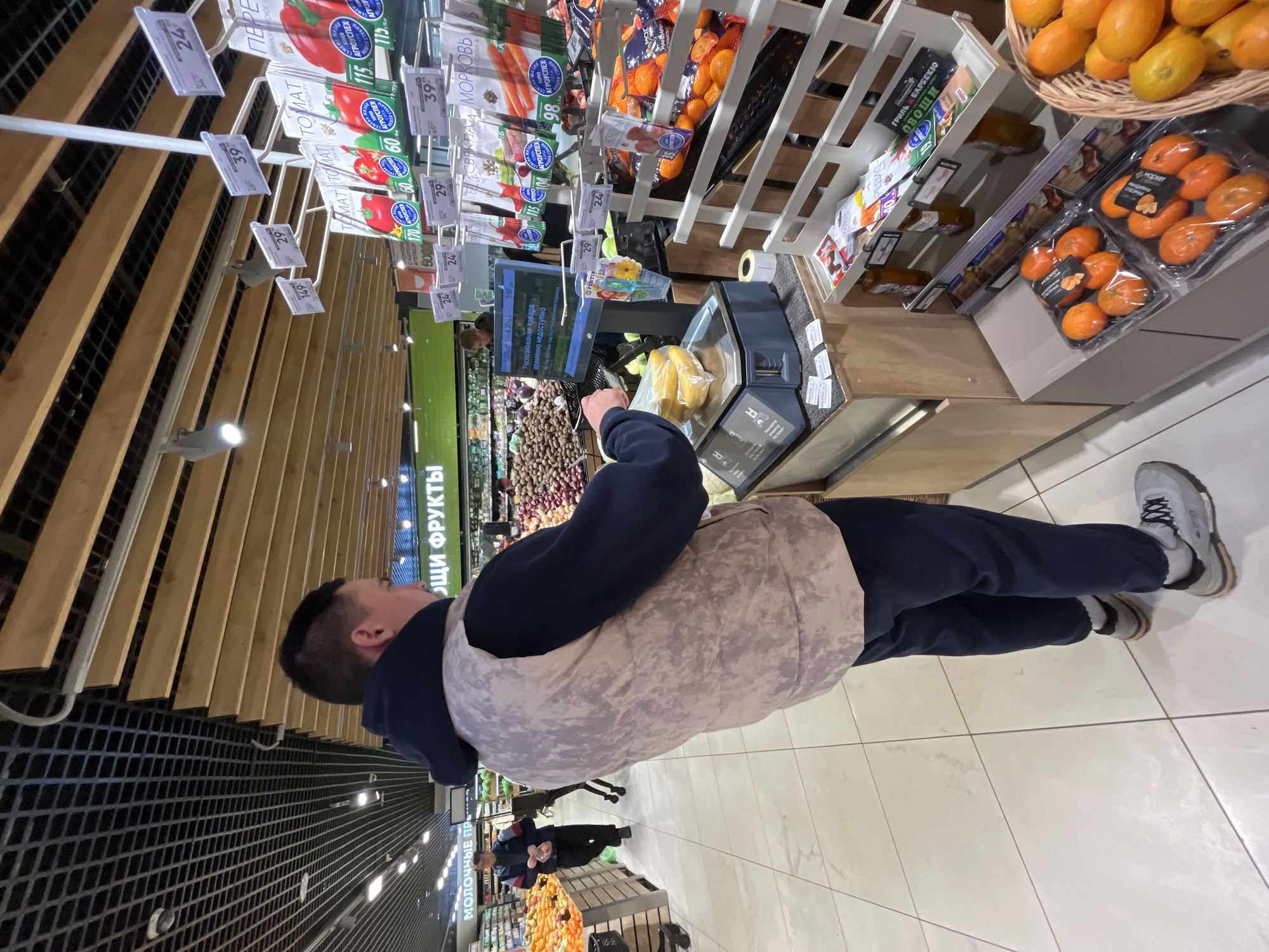 A person in a navy blue top and light beige vest is using a self-checkout kiosk at a supermarket. The kiosk displays a message in Russian stating "Product recognition temporarily unavailable". Items, possibly bananas, sit on the scale of the kiosk. Around the kiosk are various produce items, including tomatoes, carrots, mandarins, and potatoes, with price tags in Russian. The supermarket has a modern interior, with a grid-patterned ceiling and wooden accents. The location is inferred to be in Moscow, Russia, based on the Cyrillic text on the signage and products.