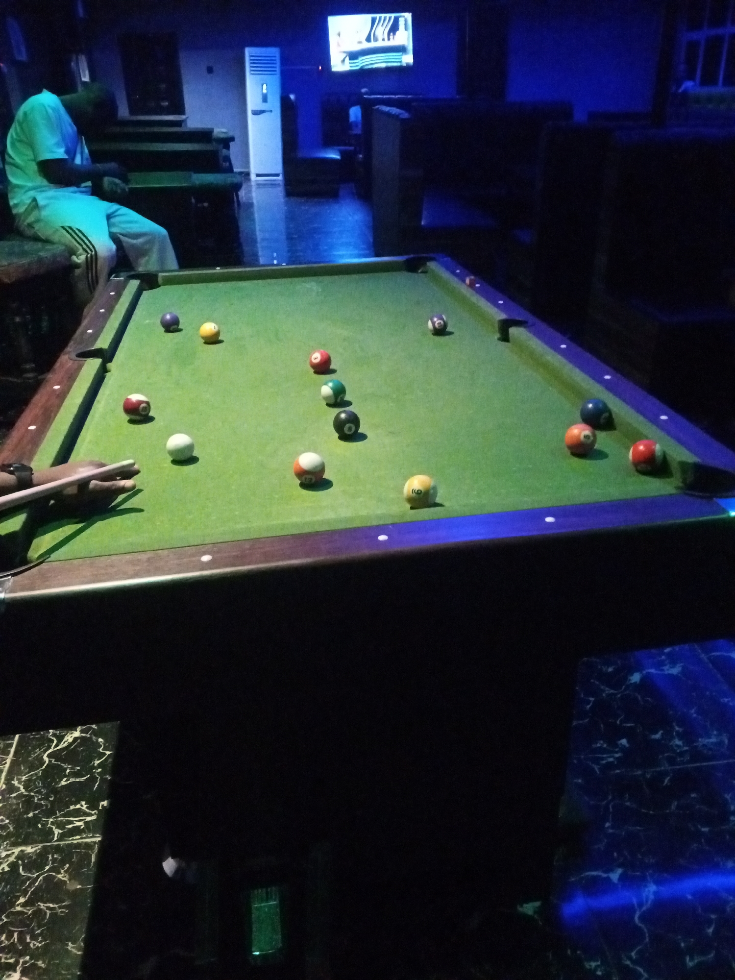 A man wearing a light-colored shirt and trousers sits on a bench to the left of a pool table. The pool table is green with a wooden frame and is set up for a game with several colored balls scattered across its surface. In the background, a tall white air conditioning unit is visible, as well as dark seating and a television screen displaying an image. The scene appears to be indoors, possibly in a bar or recreational center. The overall lighting is dim with blue accents in the environment.