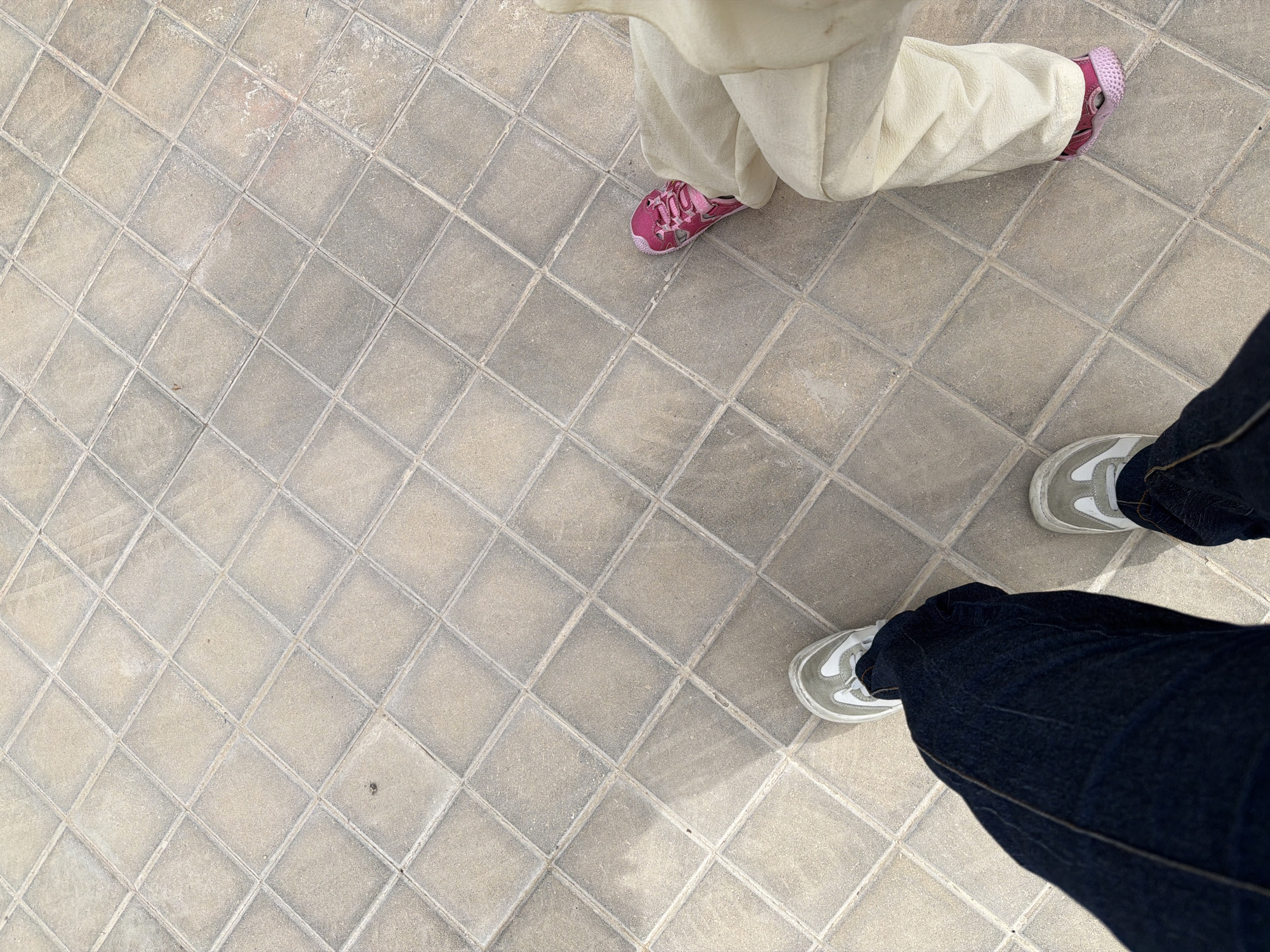 This image is a high-angle, close-up shot of two pairs of feet standing on a tiled patio. The tiles are square and laid in a diamond pattern, with light grout lines. The lighting suggests it is daytime, with shadows cast across the tiles, indicating the sun is at an angle.
One pair of feet is wearing dark denim jeans and white and gray sneakers. The other pair of feet is wearing loose, light-colored pants and a pair of pink, child-sized sneakers with star-shaped embellishments. The presence of the smaller sneakers suggests a child is present, possibly walking with an adult.
The scene is casual and appears to be outdoors, given the sunlight and outdoor tiling. There is no visible text in the image. The overall mood is relaxed and everyday, capturing a moment of people standing on a tiled surface.