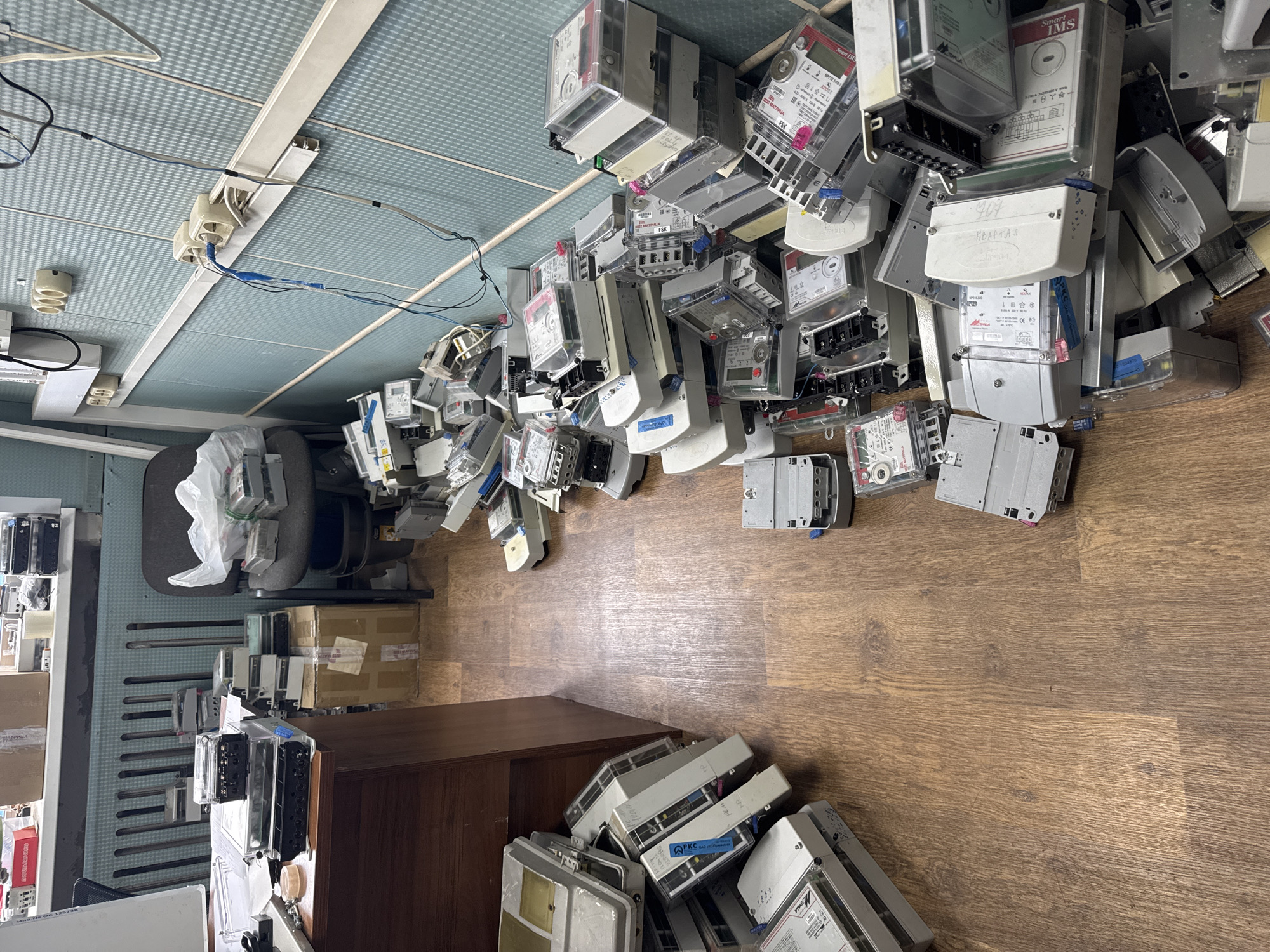 The image depicts an interior space in Chaikovsky, Russia, cluttered with a large quantity of electric meters scattered across a wooden floor. Many meters are piled near a dark wooden cabinet, with a denser accumulation towards the center of the room, extending toward a grey office chair and a desk with additional meters and equipment. The ceiling is a pale blue with visible light fixtures and electrical wiring. The room has a utilitarian, disorganized appearance suggesting a storage or workshop area. There are no people visible.