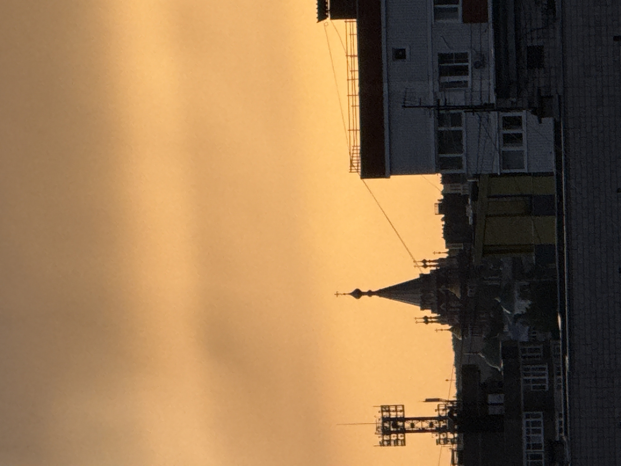The image shows an urban scene, likely in Izhevsk, Russia, at twilight or sunrise. The sky is an intense orange. The skyline features a church spire and Soviet-era high-rise buildings, one of which has a visible external ladder. An electrical grid or antenna is in the foreground. The buildings and structures appear dark in silhouette against the bright sky. The light suggests either smoke or extreme atmospheric conditions. A brick building also occupies a small section of the upper right corner.