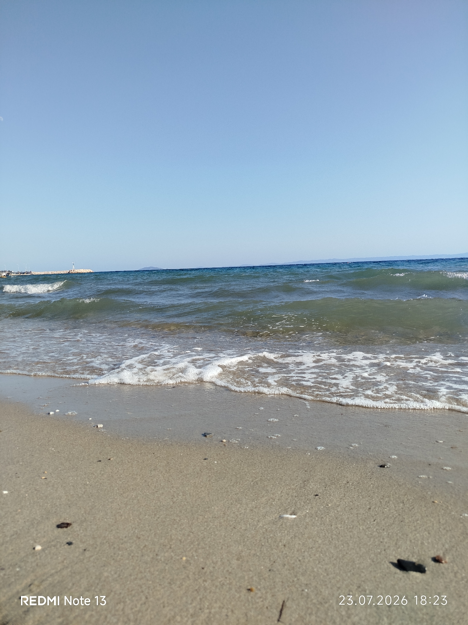 The image depicts a serene beach scene with the calm sea gently lapping at the shore. The sand is a light beige color and appears clean, with scattered small pebbles and shells. The water is a clear blue-green, with small waves creating white foam as they break on the sand. The horizon shows a clear blue sky with a faint haze. In the distance, a breakwater extends into the sea, topped with a small white structure, possibly a lighthouse or a marker. The overall environment suggests a tranquil coastal setting. The location is identified as Sithonia Municipal Unit, Greece, likely a seaside town or village within this region. The image also contains overlaid text indicating "REDMI Note 13" and a date "23.07.2026 18:23", which are likely camera metadata and not part of the scene itself. There are no people visible in the image.