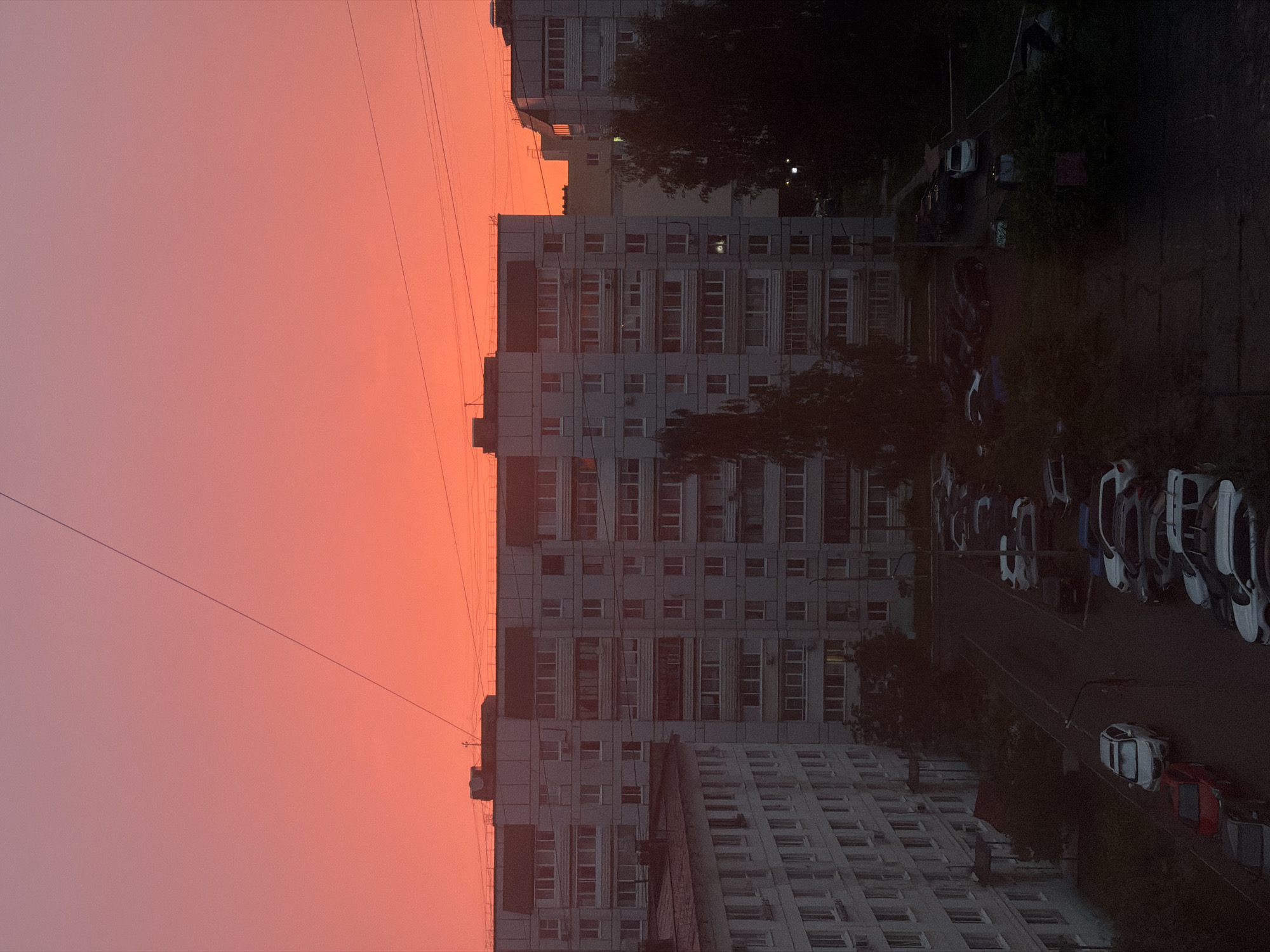 The image captures a twilight scene in Chaikovsky, Russia, characterized by a vibrant, gradient sky transitioning from deep orange near the horizon to a lighter pinkish hue above. Multiple apartment buildings, typical of Soviet-era architecture, dominate the midground. These multi-story structures are uniformly gray or light-colored, with numerous windows arranged in a grid pattern. Some windows show faint interior lights. In the foreground and midground, parked cars are visible in a courtyard or parking lot. The cars are of various makes and models, predominantly in dark colors, with a few lighter-colored vehicles. Trees with dense foliage are scattered throughout the scene, particularly towards the right and left edges, some casting shadows. Overhead wires crisscross the sky. The overall atmosphere is one of an urban residential area at dusk, with the dramatic sky providing a striking backdrop to the mundane, everyday elements of the cityscape. There are no people visible in the image.