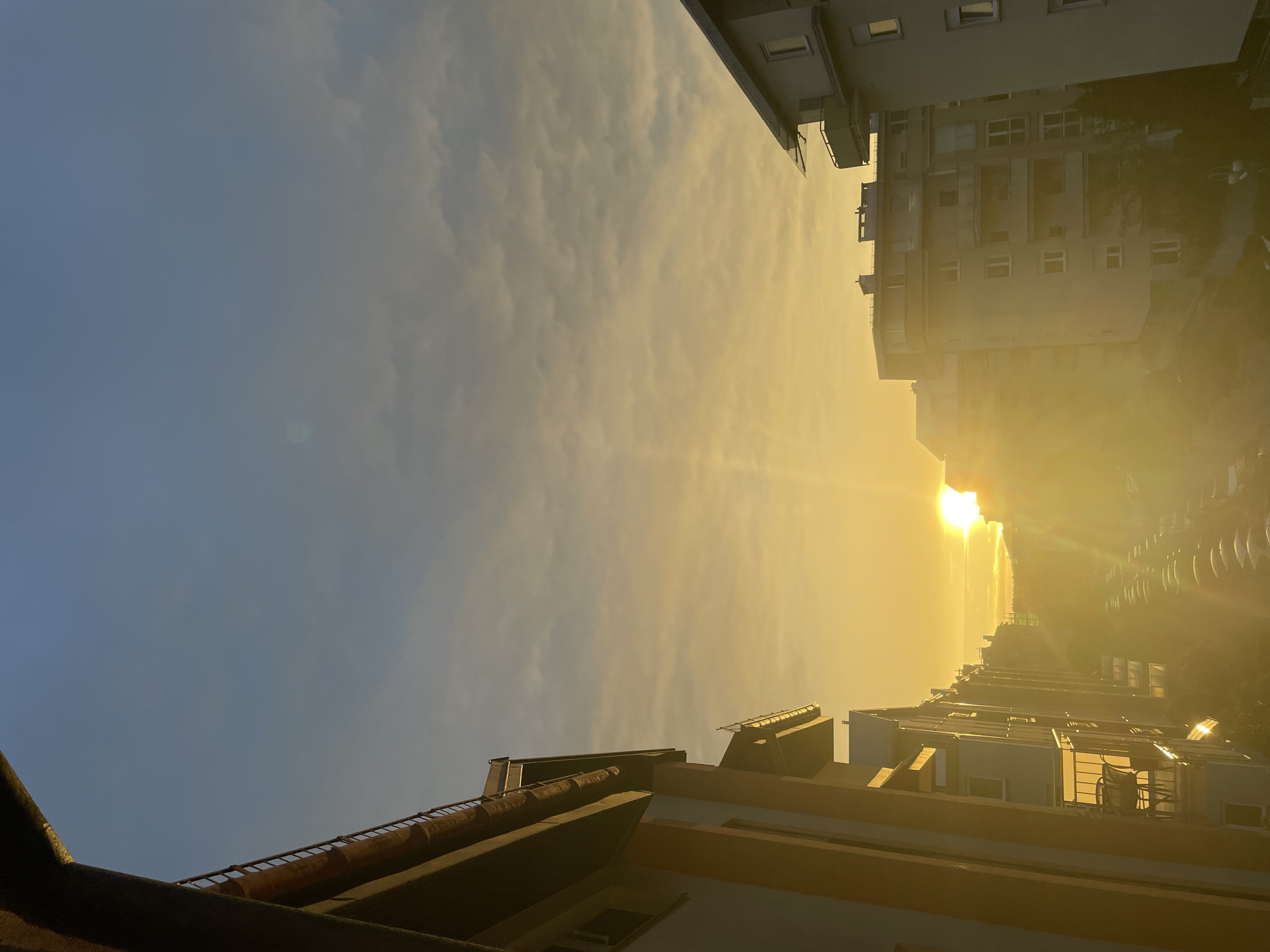 The image depicts an urban or residential street scene at either sunrise or sunset, viewed from an elevated position, likely from a window or balcony of a building. The location within the city of Unknown, Unknown cannot be determined from the visual information.
The sky in the upper portion of the frame is a clear, light blue, transitioning to a yellowish-orange hue with scattered, thin, altocumulus-like clouds towards the horizon. The sun is prominently visible low on the horizon, slightly to the right of the central vertical axis, emitting intense golden light and significant lens flare, particularly in the lower half of the image.
On the left side, the immediate foreground shows a dark, architectural overhang and a railing, suggesting the vantage point is from a building. Beyond this, a row of multi-story residential buildings, predominantly light-colored with dark roofs, extends into the distance. These buildings feature multiple windows and balconies, some of which show outdoor furniture. The facades of these buildings are brightly illuminated by the direct sunlight.
On the right side, another multi-story, light-colored residential building occupies a significant portion of the frame. This building also displays balconies and visible windows, with its upper sections reflecting the golden light of the sun.
Between the rows of buildings, a street is visible, stretching into the background. The street appears to be lined with numerous parked cars on both sides, arranged in rows. Some trees or foliage are discernible along the street, as are what appear to be streetlights. The distant buildings along the street are obscured by the intense sunlight and atmospheric haze.
No people are visible within the frame. The overall light quality is warm and golden, characteristic of the "golden hour" of photography.