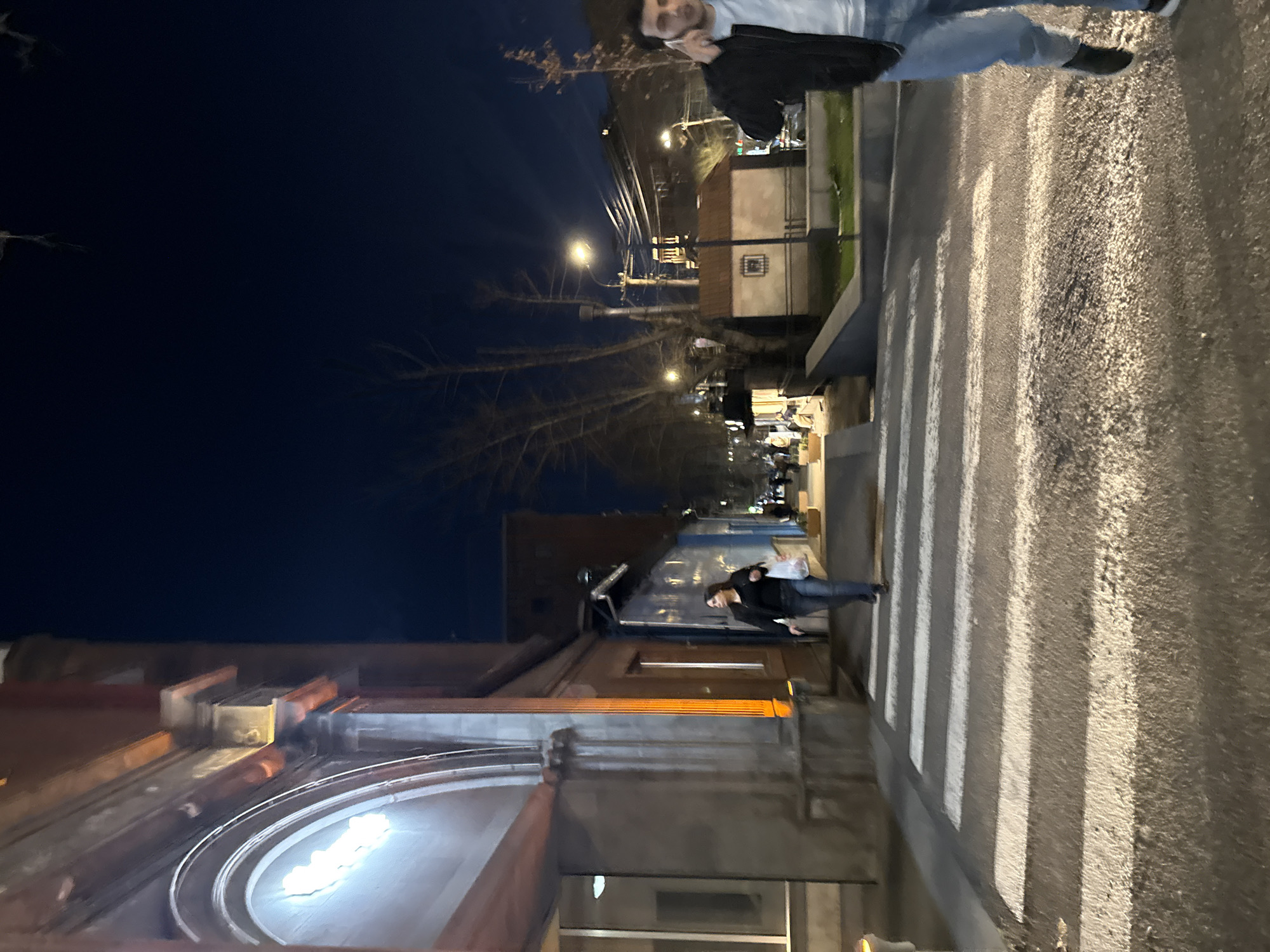The image depicts a street scene at night in Yerevan, Armenia. The foreground shows a building with an arched entrance and lit windows. A person is walking across a zebra crossing in the middle ground. The street is illuminated by streetlights. A second person is blurred in the background. The sky is dark and clear.