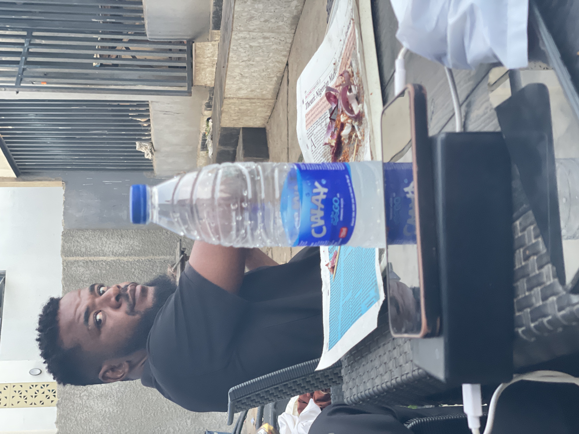 The image shows a man with dark skin and a beard, wearing a black shirt. He is seated at a table with a bottle of CWAY water. A newspaper with the headline "Theurl: Nigerian Made" is on the table, with some remnants of food (possibly onions and meat) on it. A cell phone and a black portable charger connected with a white charging cable are also on the table. The table is a wicker type with a black laptop and a white cable on it. Outside the enclosed area is a grey wall with the top part being an open iron grate. The location is identified as Akwanga, Nigeria.