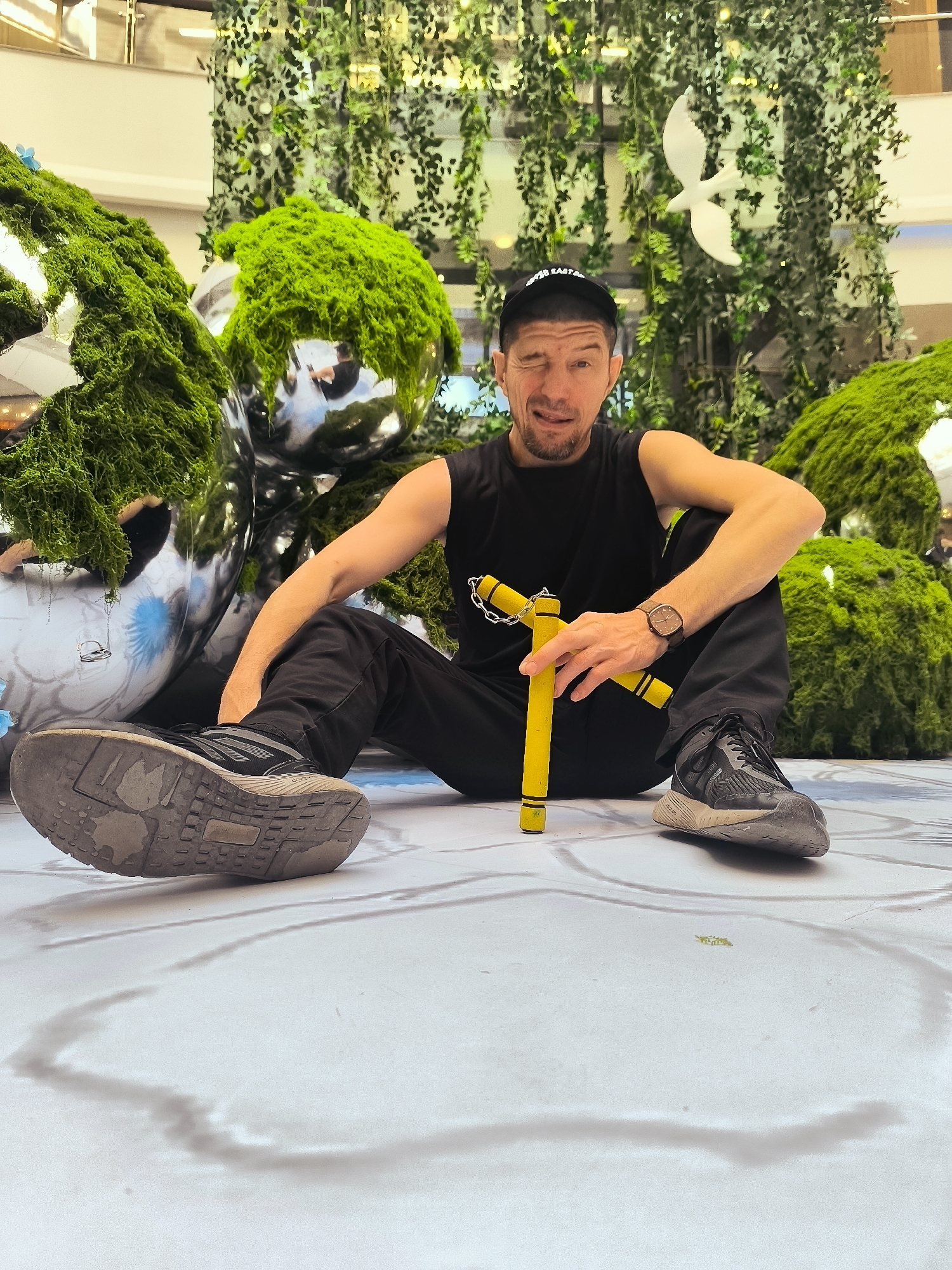 A man wearing a black t-shirt, black pants, a black baseball cap, and black sneakers sits on a white floor with a pattern. He is holding a yellow nunchaku in his right hand. He is winking at the camera. Behind him, there are large spherical decorations covered in green moss, and a wall covered in hanging green vines with a white bird decoration in the center. The scene appears to be an indoor public space, possibly a mall or exhibition.