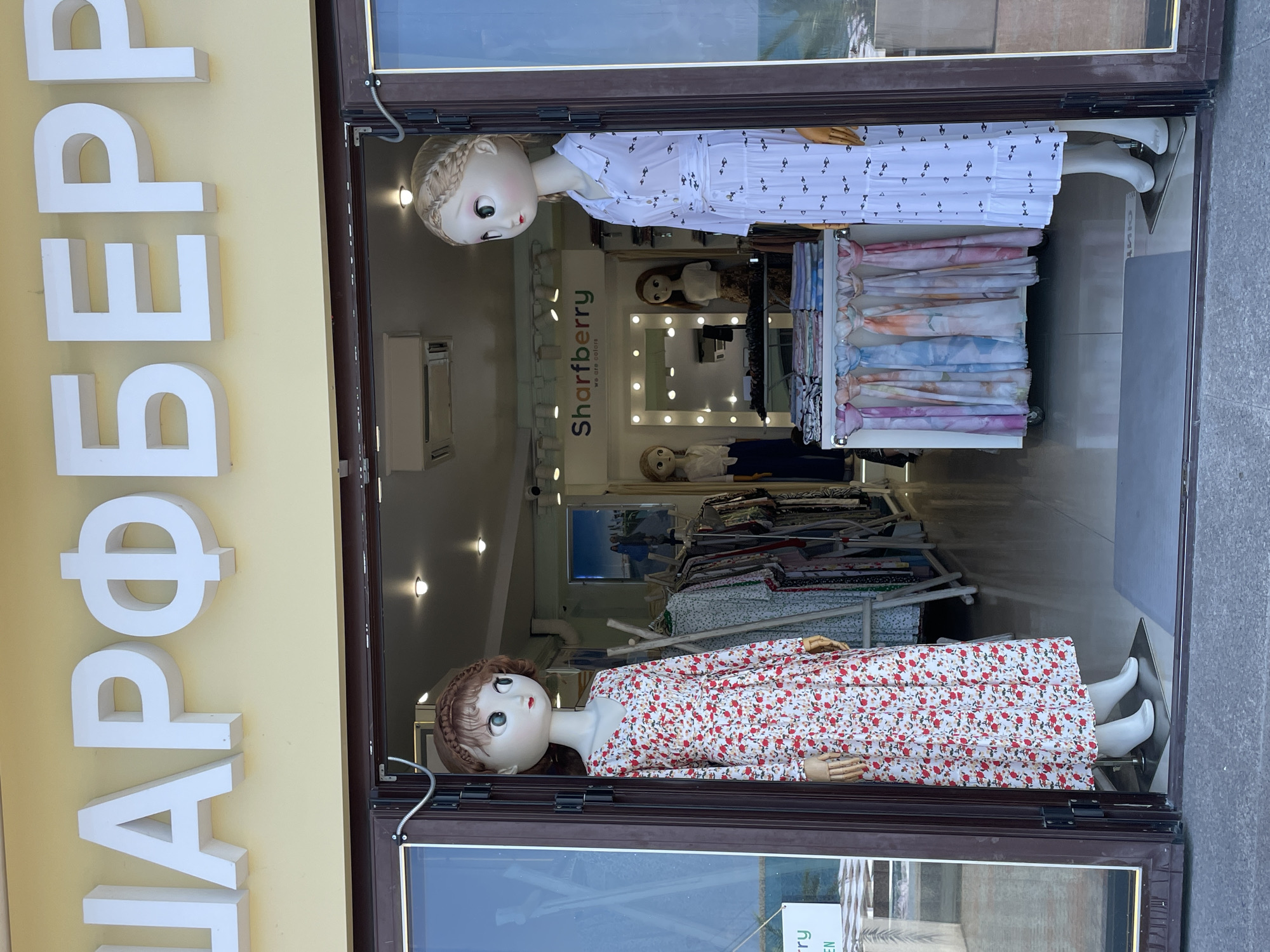 This is a medium shot, captured in daylight, of a shop window display. The window is divided into two vertical sections by a dark brown frame. In the left section of the window, large, white, three-dimensional letters are mounted vertically on a pale yellow wall, spelling out "MAPPER." This likely refers to the shop's name.
The right section of the window displays mannequins dressed in clothing and a rack of colorful fabrics. The upper part of this section features a mannequin with fair skin, blonde braided hair, and large, prominent eyes. It is wearing a white dress with a black, repeating pattern of small fish. The mannequin's right arm is bent, resting on its chest.
Below this, a sign with green and white text reads "Sharberry we are colors." The sign is illuminated by a light fitting. Further back in the shop, more mannequins are visible, as well as racks of clothing and fabric bolts.
In the lower part of the right window section, another mannequin is displayed. This mannequin also has fair skin and braided blonde hair, with large eyes. It is wearing a long-sleeved dress with a white background and a repeating pattern of small red flowers. Its hands are resting by its side, and its white shoes are visible.
A rack of folded fabrics, in various pastel colors, is positioned in front of the lower mannequin. The shop appears to be well-lit, with spotlights visible. The overall impression is of a clothing store showcasing its merchandise through window displays. The location is indicated as Sochi, Russia, and the time of day is daytime.