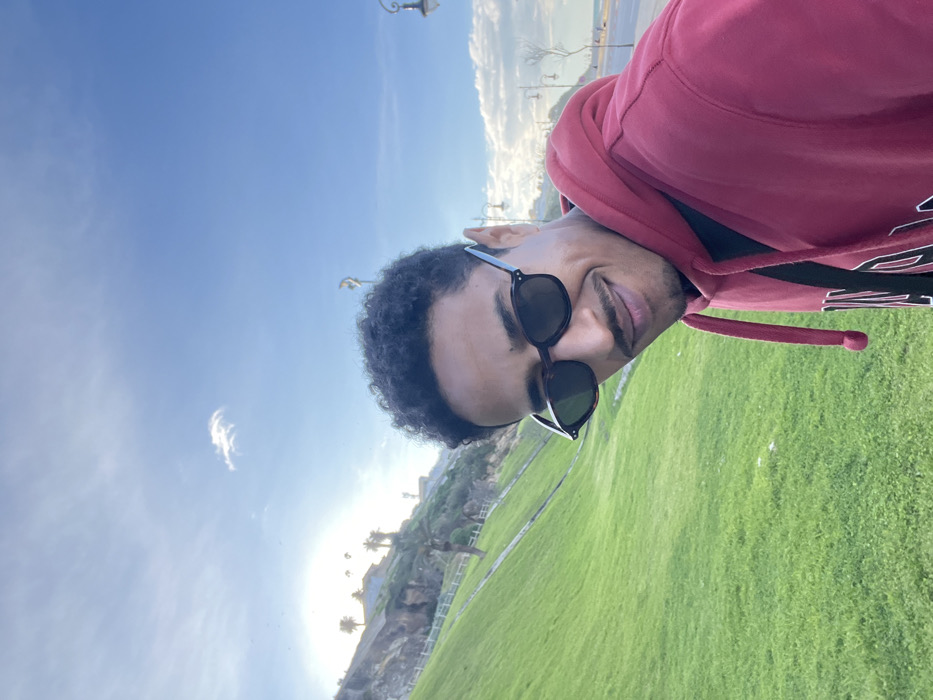 A male individual with dark, curly hair and a mustache is depicted in a selfie-style photograph, smiling and looking slightly away from the camera's direct axis. He is wearing dark, tortoiseshell-framed sunglasses and a red hooded sweatshirt. The scene is an outdoor coastal environment in Tanger, Morocco. To the subject's left, a steep, manicured green grassy hillside ascends, featuring a white fence, scattered palm trees, and distant white buildings near its summit, strongly illuminated by the low sun. To the subject's right, a paved promenade equipped with ornate streetlights borders a sandy beach and the sea. The sky is clear blue with minimal white cloud cover, and the sun is visible low on the left, casting a bright glare.