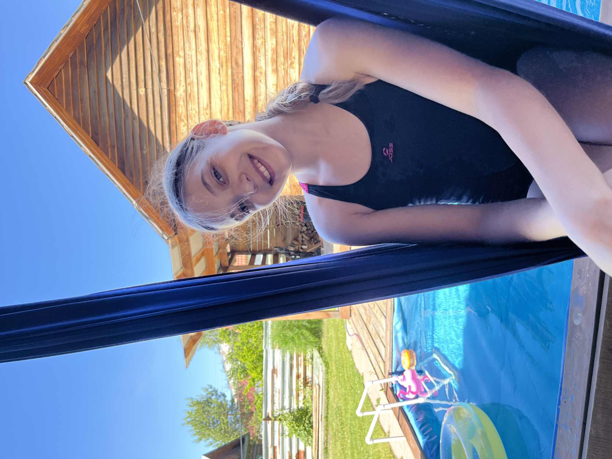 A young girl with blonde hair in a braid is smiling at the camera, seated in a dark blue hammock. She is wearing a black swimsuit with pink trim and a "JOSS" logo. Behind her, a wooden structure with a sloped roof is visible. To the left, a blue inflatable pool with a yellow ring float and two pink flamingo floats is in a backyard with green grass and a white metal fence. The sky is clear and blue.