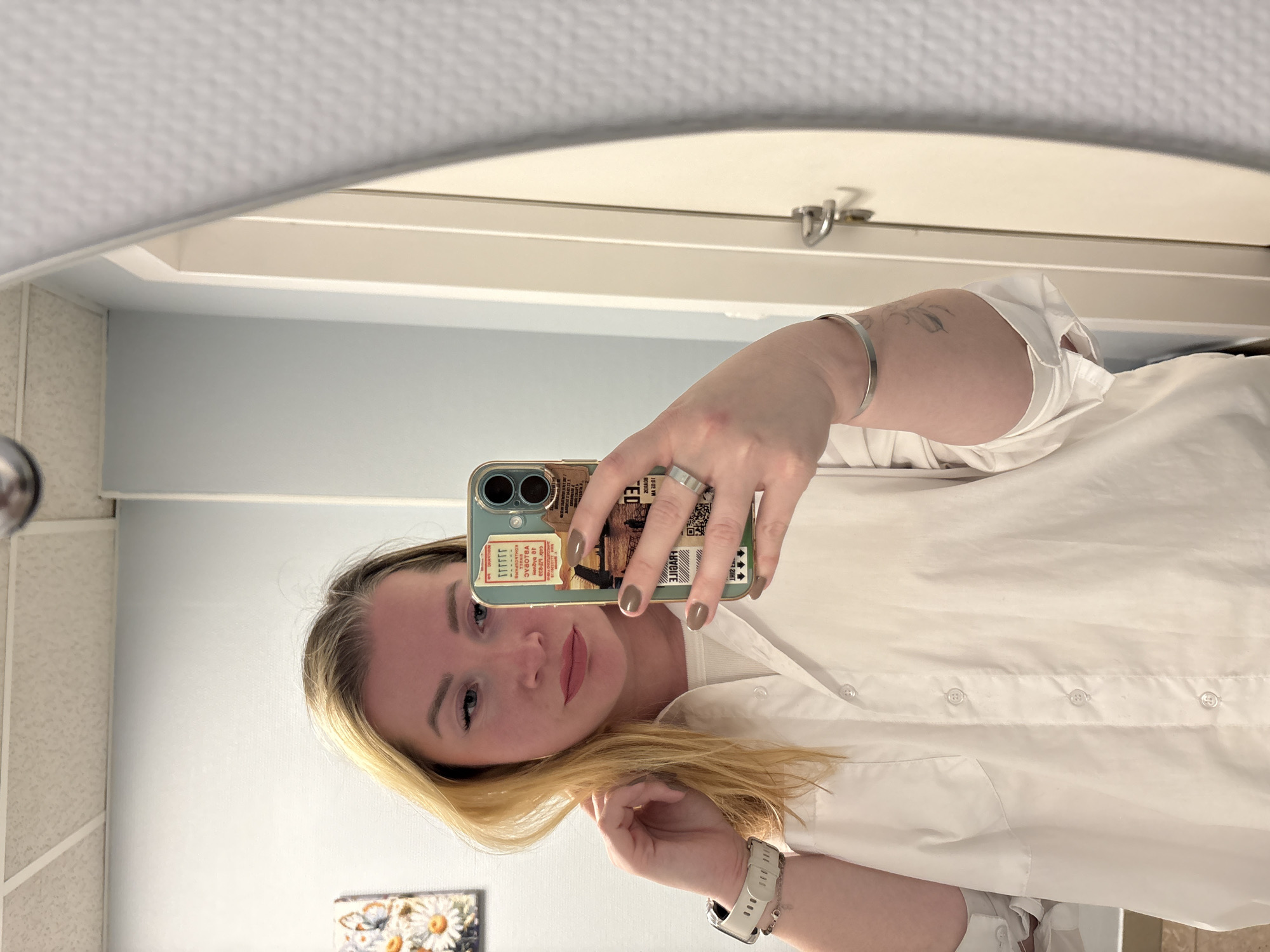 This image is a mirror selfie taken by a woman indoors, presented rotated 90 degrees clockwise. The subject is a woman with blonde hair, looking towards the camera with a neutral expression. She is wearing a white button-down shirt. On her right arm, she wears a simple silver bangle bracelet, and a small, delicate tattoo of a leafy design is visible on her inner forearm. On her left hand, she wears a silver band ring on her ring finger and a smartwatch with a white band on her wrist. Her fingernails are painted a muted, light taupe or greige color.
She holds a light blue/teal smartphone with a distinctive, multi-patterned case in her left hand, obscuring parts of her face. The phone case features several rectangular designs that resemble tickets or stamps, some with faded or stylized text. Partially visible text on the case includes reversed words like "VVORBGA" and "ERBICE", along with "0.01" and what appears to be an illustration of a bridge or structure.
The background reflected in the mirror reveals an indoor setting, likely a bathroom or dressing area. A light blue wall is visible, and to the far left, a tiled or textured wall. At the bottom-left corner of the mirror, a small framed picture depicting daisies and butterflies can be seen. Above the woman (if the image were rotated upright), the reflection shows a white ceiling with a textured surface, a recessed light fixture, and a white door or cabinet frame with a silver latch or hinge.
The activity is a self-portrait taken using a mirror. The time of day is indeterminate due to indoor lighting. The location context provided is Veliky Novgorod, Russia.