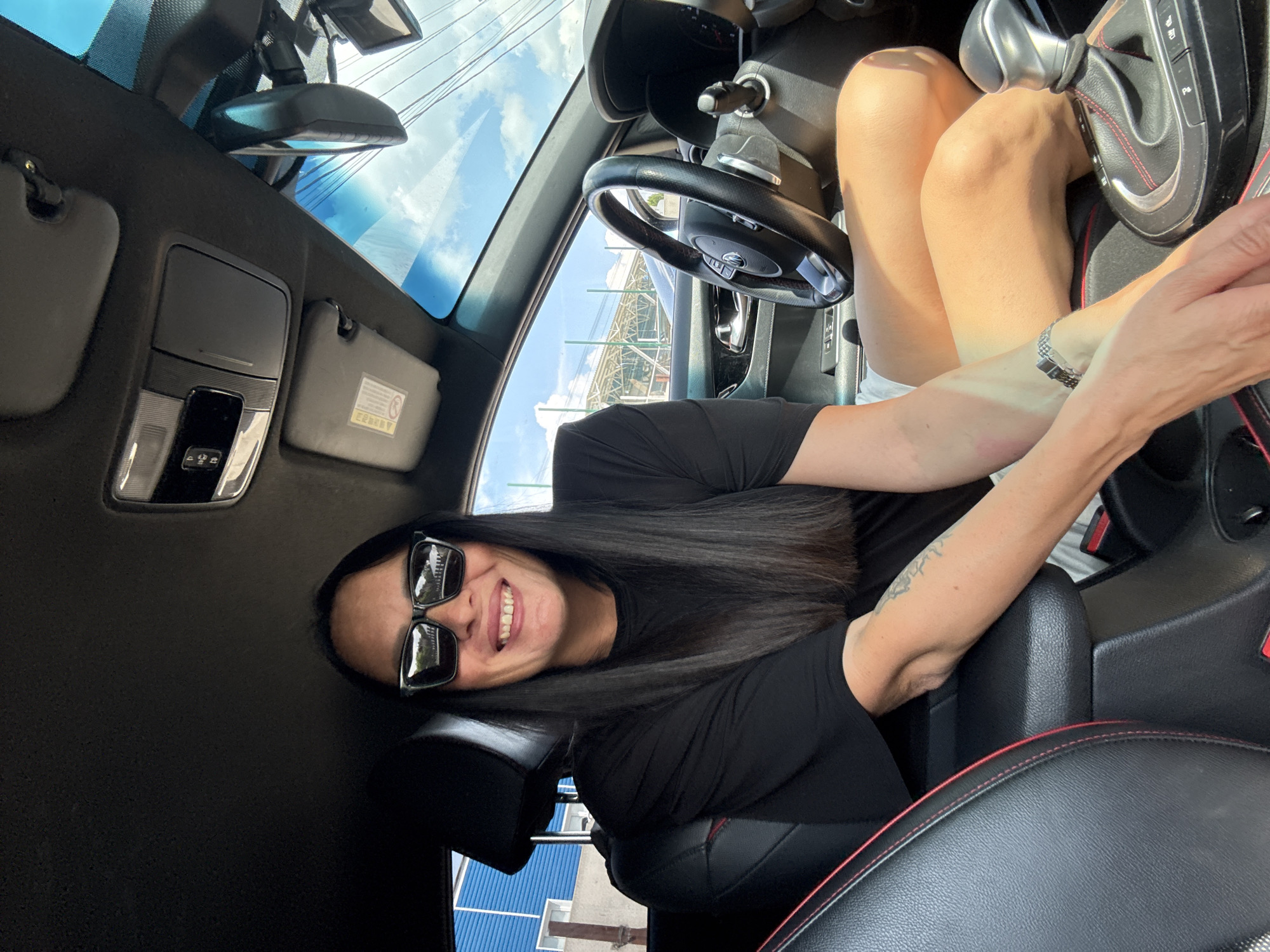 A woman with long, dark hair and sunglasses is sitting in the driver's seat of a car. She is smiling and wearing a black t-shirt and white shorts. Her arms are resting on the center console. The interior of the car is visible, including the steering wheel and gear shift. Outside the car, a blue building and a fence can be seen. The sky is blue with some clouds. The image does not provide any information to place the location in Chaikovsky, Russia.