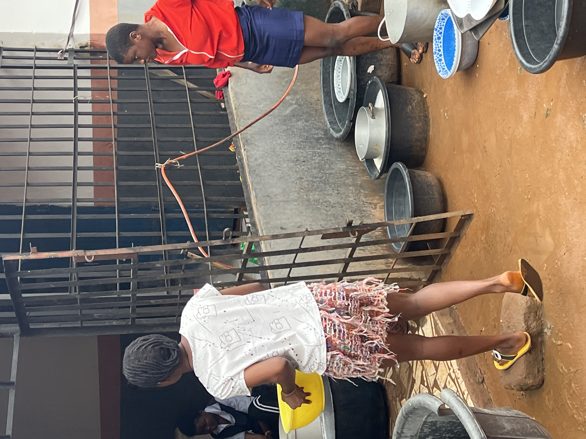 In Nkpor, Nigeria, two young women are engaged in an outdoor activity near a gated structure. The woman on the left, wearing a white t-shirt with cartoon characters and a multi-colored, fringed skirt, holds a large yellow bowl. She is facing away from the camera and has her hair styled in numerous braids. The woman on the right is wearing a red hooded jacket and a dark blue skirt. She is looking to her left. The environment is a dirt ground, wet in places, with numerous large black plastic containers filled with water and other items, including metal pots and a blue and white patterned bowl. A metal gate with vertical bars runs across the background, partially obscuring a building with orange and white striped elements. A red hose is attached to the gate. The scene appears to be a communal area, possibly for washing or food preparation, given the presence of the containers and the people's activities.