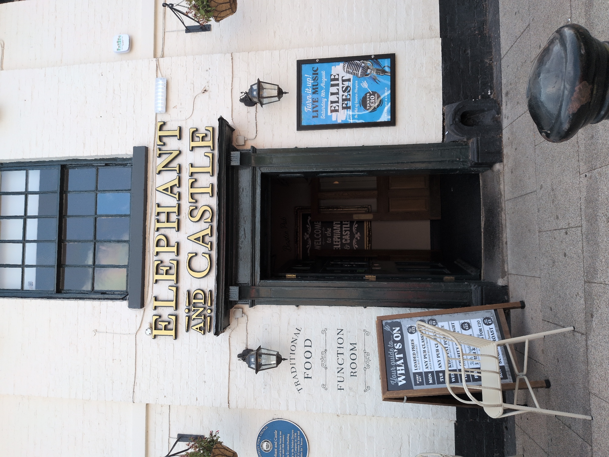 The image shows the exterior of the Elephant and Castle pub in Dawley, United Kingdom. The pub has a white brick facade with large gold lettering spelling out "ELEPHANT AND CASTLE" above a dark green doorway. To the left of the door, there are signs indicating "TRADITIONAL FOOD" and "FUNCTION ROOM." A chalkboard sign titled "Your guide to... WHAT'S ON" is placed in front of the entrance, displaying various daily specials. To the right of the doorway, a blue poster advertises "LIVE MUSIC" for "ELLIE FEST" on Saturday, 8th August, in aid of Severn Hospice, with tickets costing £10 per person. A vintage microphone graphic is featured on the poster. Above the door, a black framed window with multiple panes is visible. Two traditional-style lanterns are mounted on the wall, one to the left and one to the right of the entrance. Hanging baskets with plants are present on either side of the building. A white metal chair with an open back is positioned in front of the chalkboard sign. The pavement in front of the pub is made of grey stone slabs. A black bollard with a rounded top is visible in the lower right corner of the frame. The overall scene depicts a street-level view of a pub entrance with promotional materials and street furniture.