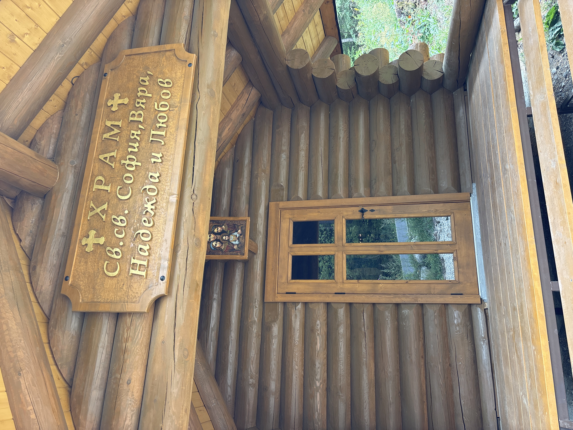 The image shows the facade of a small wooden log church, likely a chapel or hermitage, in Bansko, Bulgaria. A wooden sign above the entrance reads "ХРАМ СВ.СВ. СОФИЯ, ВЯРА, НАДЕЖДА И ЛЮБОВ" in gold Cyrillic lettering, translating to "Church of Saints Sophia, Faith, Hope, and Love". Below the sign, a small icon depicting the Virgin Mary and Child is mounted on the log wall. A wooden door with glass panes, divided into four sections, is centrally located. The building has a covered porch with wooden plank flooring and a wooden bench. The surrounding environment appears to be wooded and natural.