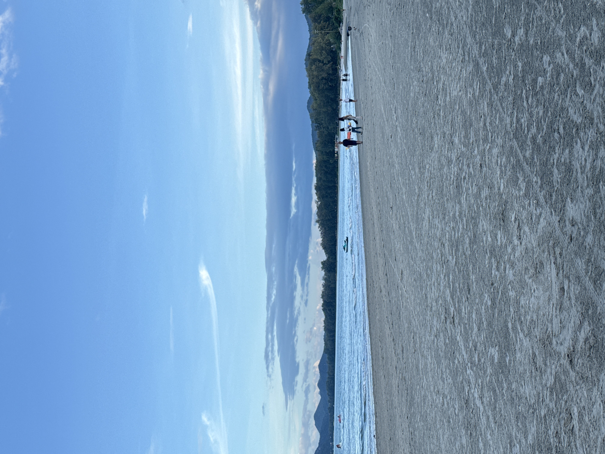 The image depicts a serene beach scene in Hua Hin City Municipality, Thailand. The foreground is dominated by a wide expanse of sandy beach, with subtle textures and tracks suggesting recent activity. In the middle ground, the calm, blue ocean stretches out, meeting the shore with gentle waves. Several small boats, including a jet ski and what appears to be a paddleboat, are visible on the water.
Further back, a line of lush green trees forms a backdrop against a range of dark, undulating mountains under a vast, pale blue sky. The sky is adorned with wispy white clouds, some of which are elongated and stretched across the horizon.
On the beach, a small group of people can be seen strolling near the waterline. One group of three individuals, two women and a man, are walking together, while another person is further down the beach. The overall atmosphere is peaceful and tranquil, indicative of an evening setting or a quiet day by the sea.