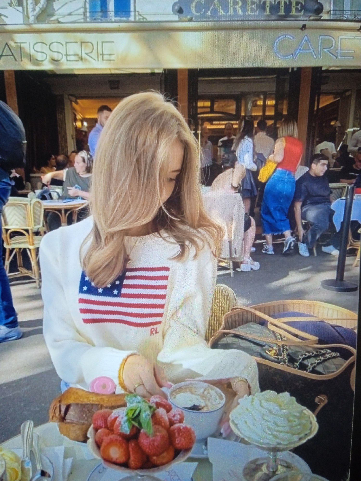This is a photograph taken outdoors in front of a cafe. The cafe has signage that appears to say "ATISSERIE" on the left and "CARETTE" on the right. In the foreground, a woman with blonde hair wearing a white sweater with an American flag design sits at a table. In front of her are several desserts, including a bowl of strawberries, a cup with a creamy dessert, and a large serving of whipped cream. A purse is visible in a basket beside her. In the background, there are other patrons sitting at tables and walking around. The overall scene suggests a casual dining experience at a cafe. The location is not Chernivtsi, Ukraine.