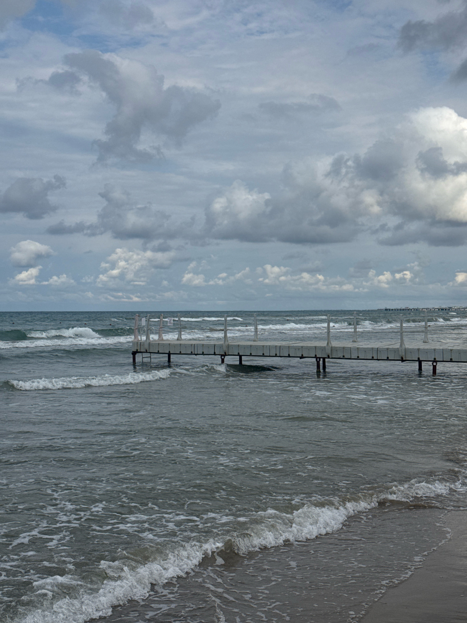 The image depicts a cloudy day at a beach in Anapa, Russia. In the foreground, gentle waves with white foam are washing ashore onto a sandy beach. A white, modular pier extends into the sea from the right side of the frame, supported by dark pilings. The sea is moderately choppy with several lines of waves. In the distance, the horizon is visible with a hazy line of structures and a few more piers stretching out into the sea. The sky is filled with dramatic, grey and white clouds, creating a somewhat overcast atmosphere. No people are clearly visible in the frame.