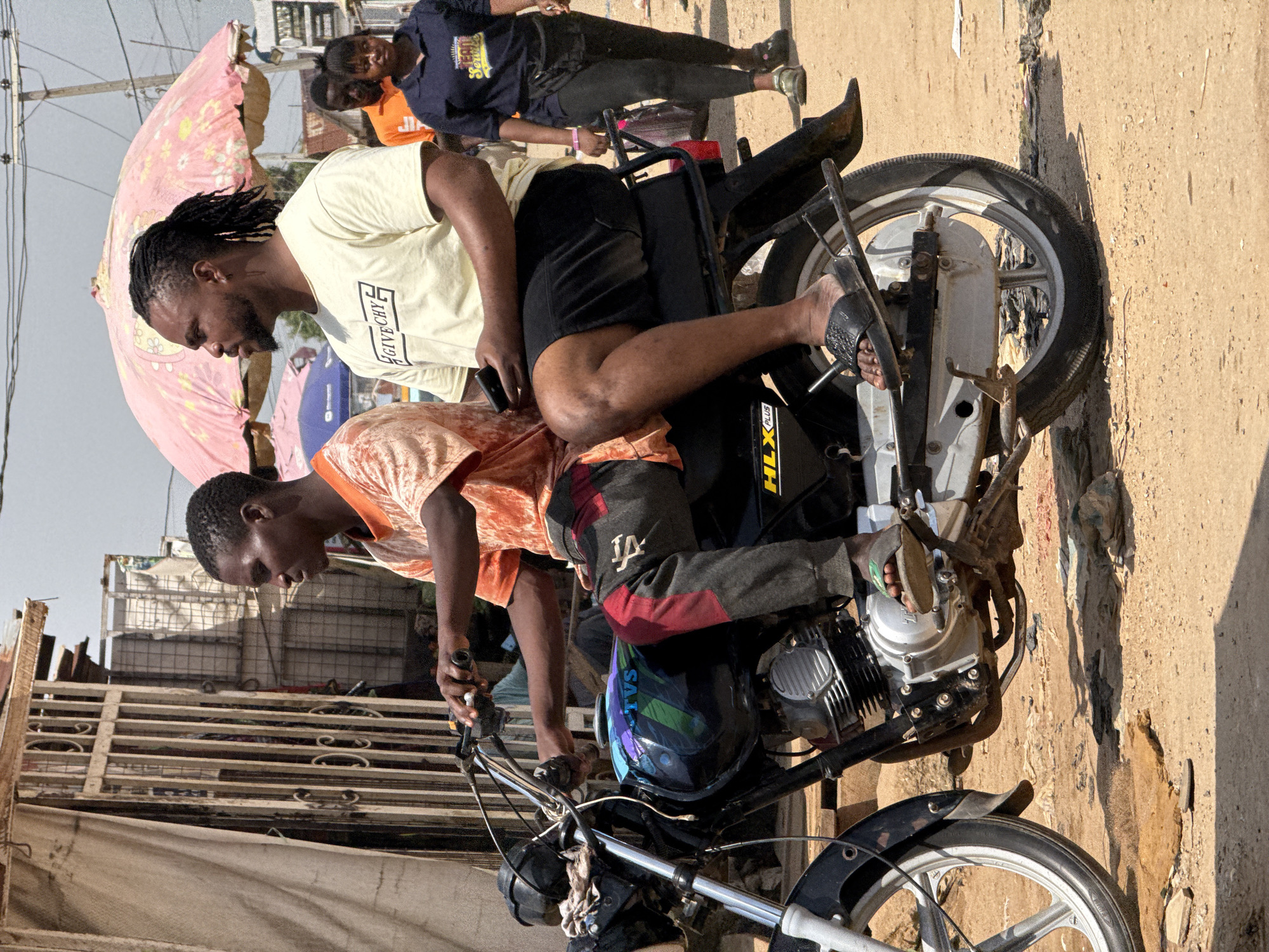 Two men are riding a motorcycle on a dirt road in Abuja, Nigeria. The man in front is driving, wearing an orange t-shirt and dark pants with red accents. The man behind him is wearing a light-colored t-shirt with "GIVE CHY" printed on it and black shorts. He is holding a black mobile phone in his right hand. The motorcycle is black with a colorful design on the fuel tank and the word "TVS" is visible. A pink umbrella is visible in the background, along with other people walking. The ground is covered in dirt and debris.