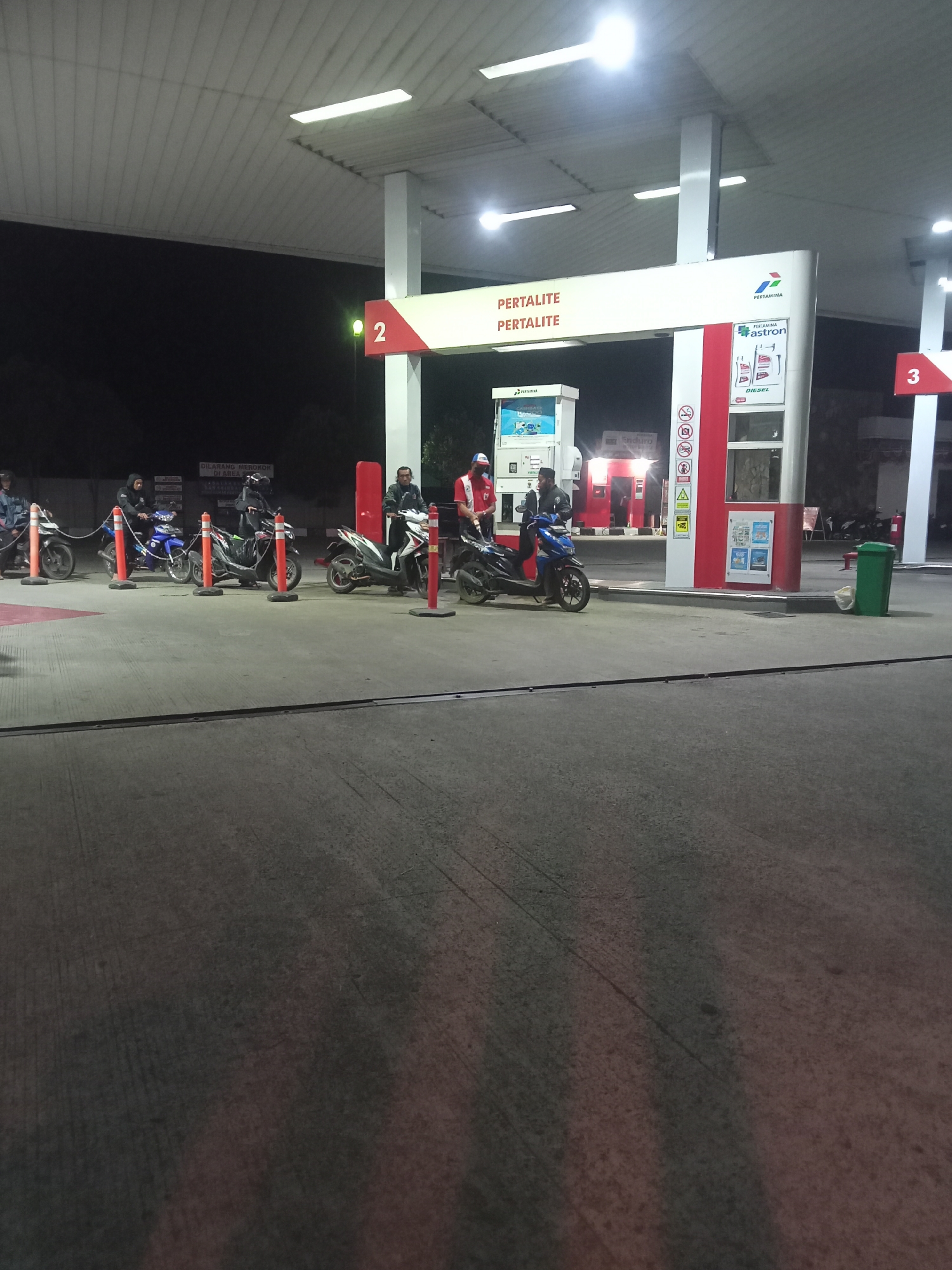 The image depicts an outdoor nighttime scene at a Pertamina gas station in Gladagsari, Indonesia. Several motorcycles are lined up at a gas pump, with riders waiting. One attendant, wearing a red uniform, stands near a motorcycle. Another attendant in a black jacket is also present. A gas pump with the number "2" prominently displayed is visible, alongside signage indicating "PERTAMINA" and fuel types like "PERTALITE" and "DIESEL". A green trash can is located to the right of the scene. The ground is paved concrete, and the area is illuminated by overhead lights. A sign prohibiting smoking is also visible.